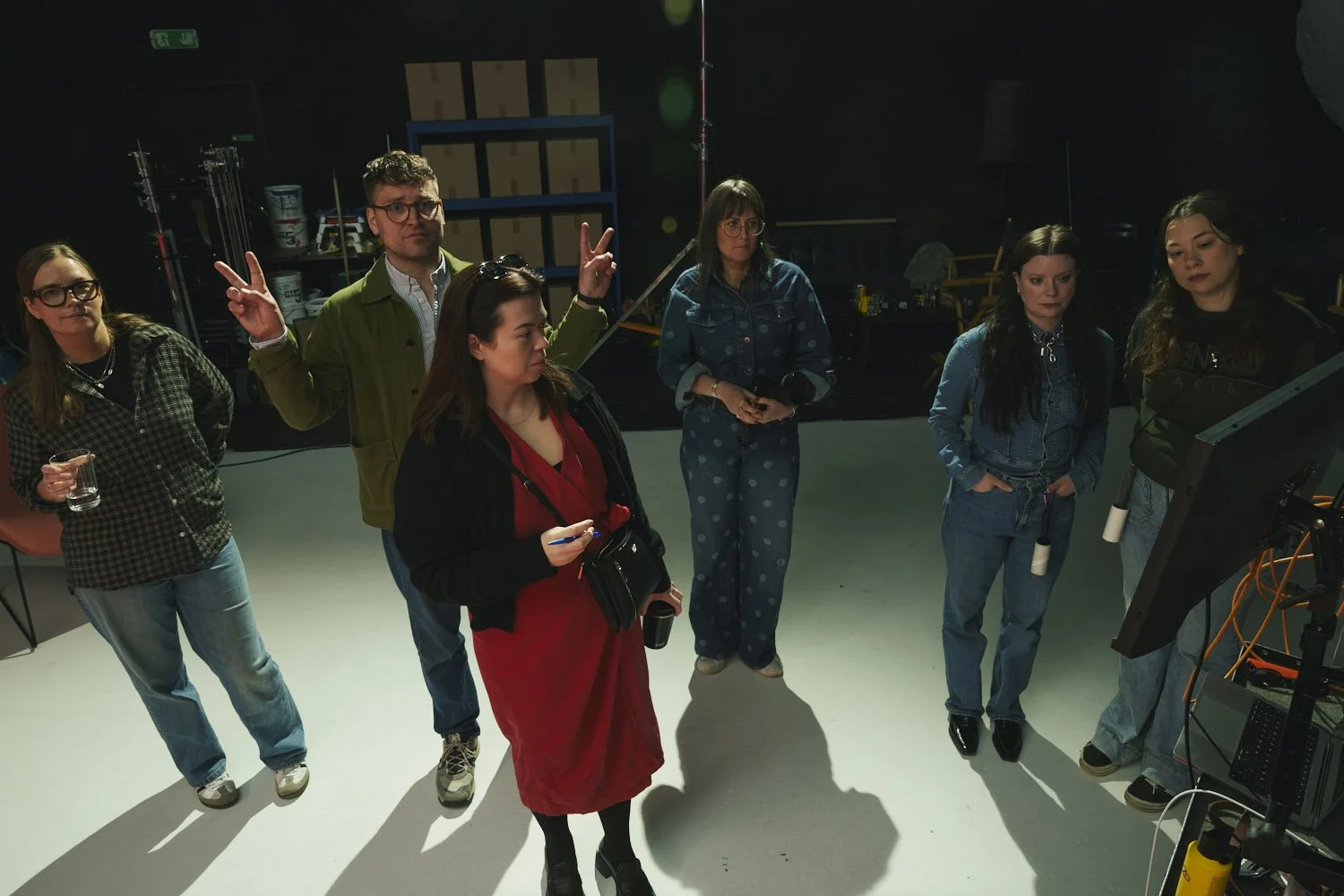 Group of six people on a film or TV set, some holding drinks or notes, standing around a monitor, with equipment and storage shelves in the background.