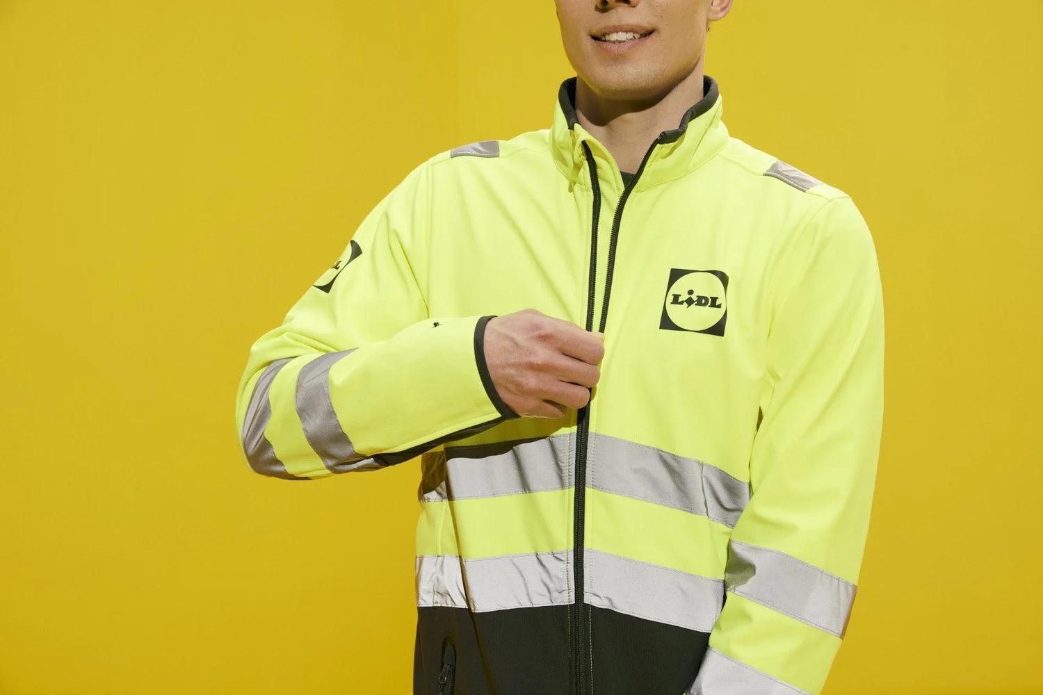 Close-up of a person wearing a high-visibility yellow safety jacket with reflective stripes and a Lidl logo on the chest, standing against a yellow background.