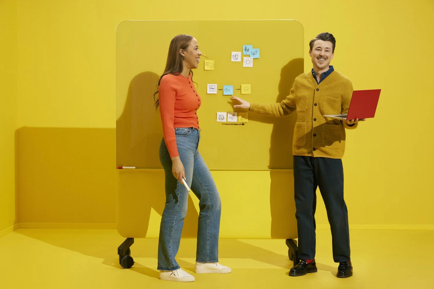 A woman and a man stand in front of a yellow wall with notes on a board. The woman is smiling, wearing a red sweater and jeans, holding a marker. The man is smiling, wearing a yellow cardigan and dark pants, holding a red laptop, and gesturing toward
