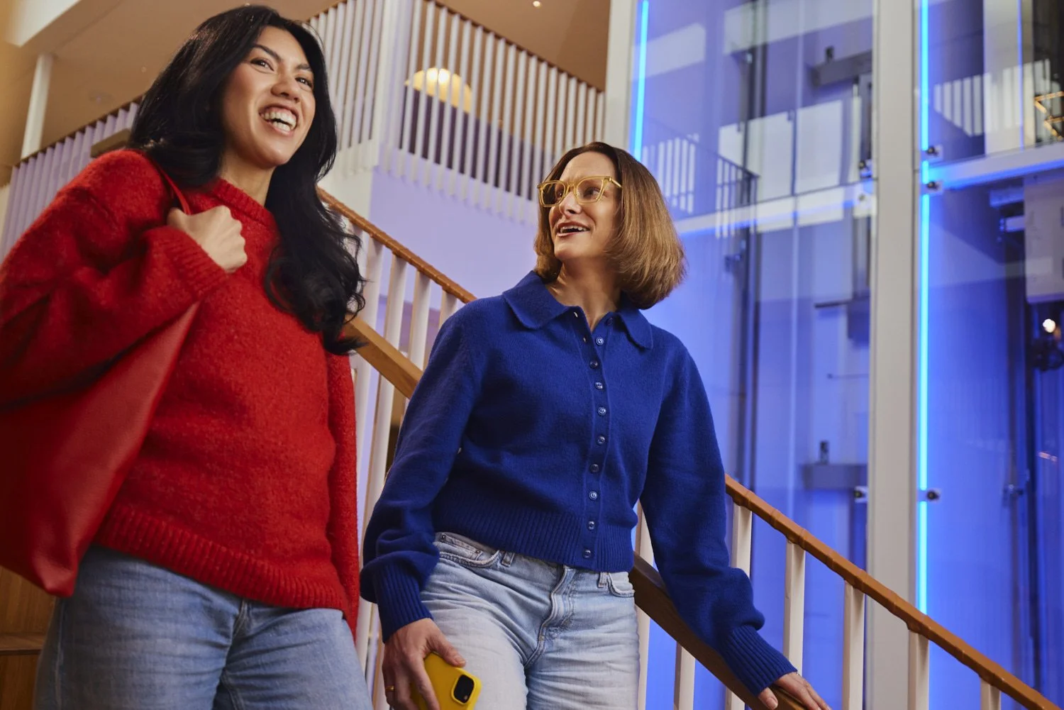 Two women talking and smiling inside a modern building with wooden stairs and glass walls illuminated by blue neon lights.