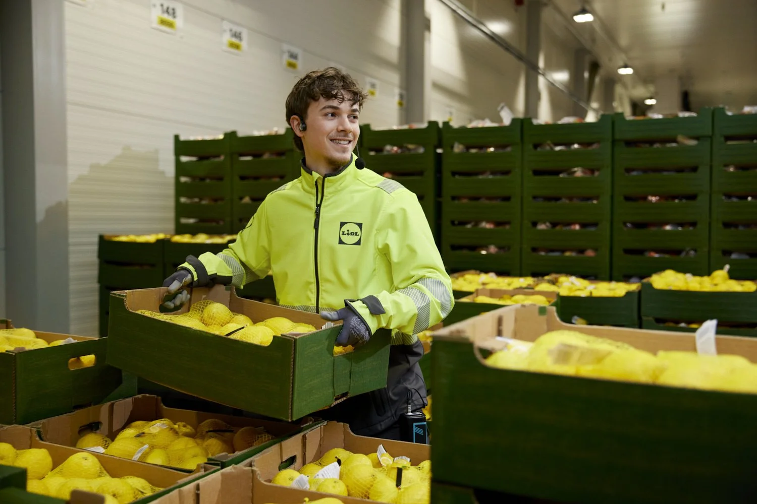 A smiling worker wearing a neon yellow jacket with the Lidl logo, handling yellow produce in a warehouse or produce section at Lidl.