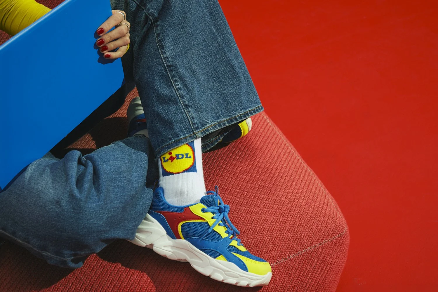 Close-up of a person's crossed legs wearing blue jeans and colorful sneakers with yellow, red, and blue accents, sitting on a red textured surface with a red background, holding a blue shopping bag.