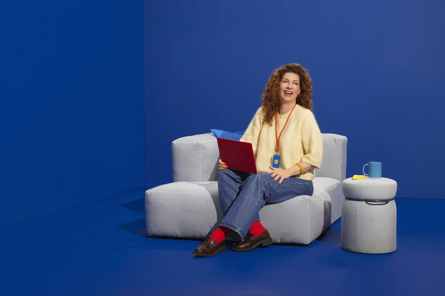 Woman with curly hair sitting on a light gray sofa against a blue background, holding a red laptop, with a blue pillow behind her, a blue mug, and a small round gray side table with a yellow remote control.