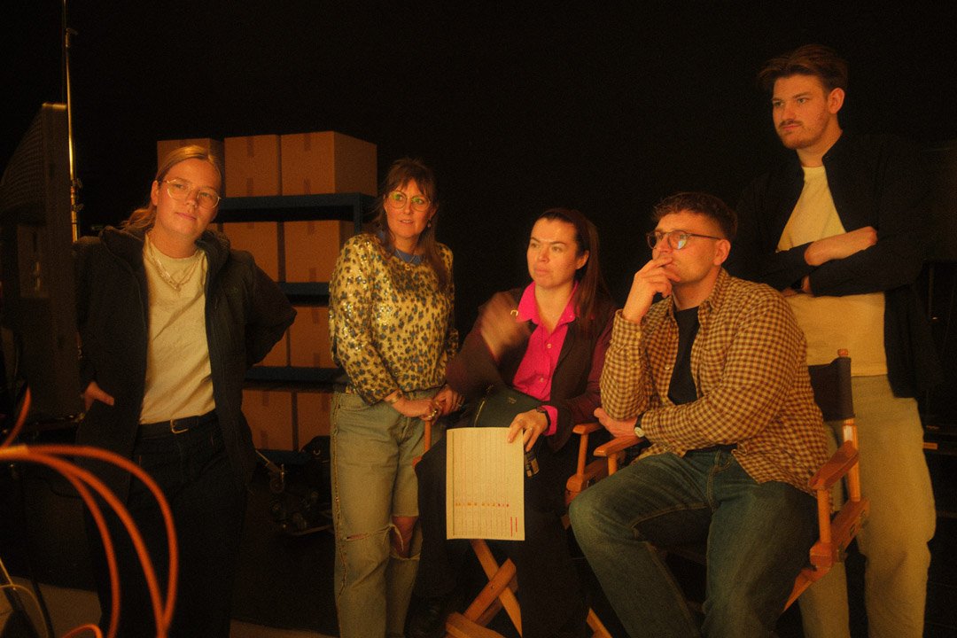 Group of five young adults in a dimly lit room, with boxes and shelves in the background, appearing to be on a film or TV set.