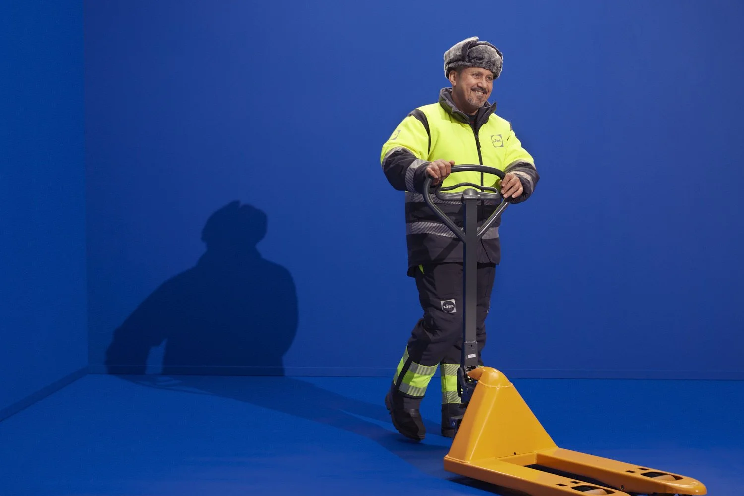 A man dressed in high-visibility work clothing and a furry hat is operating a yellow pallet jack on a solid blue background.