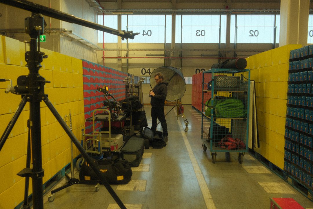 A person dressed in black with glasses is standing in a colorful warehouse or studio, holding a transparent umbrella. Surrounding them are various photography or filming equipment, including a camera on a tripod, a large light, and storage carts with