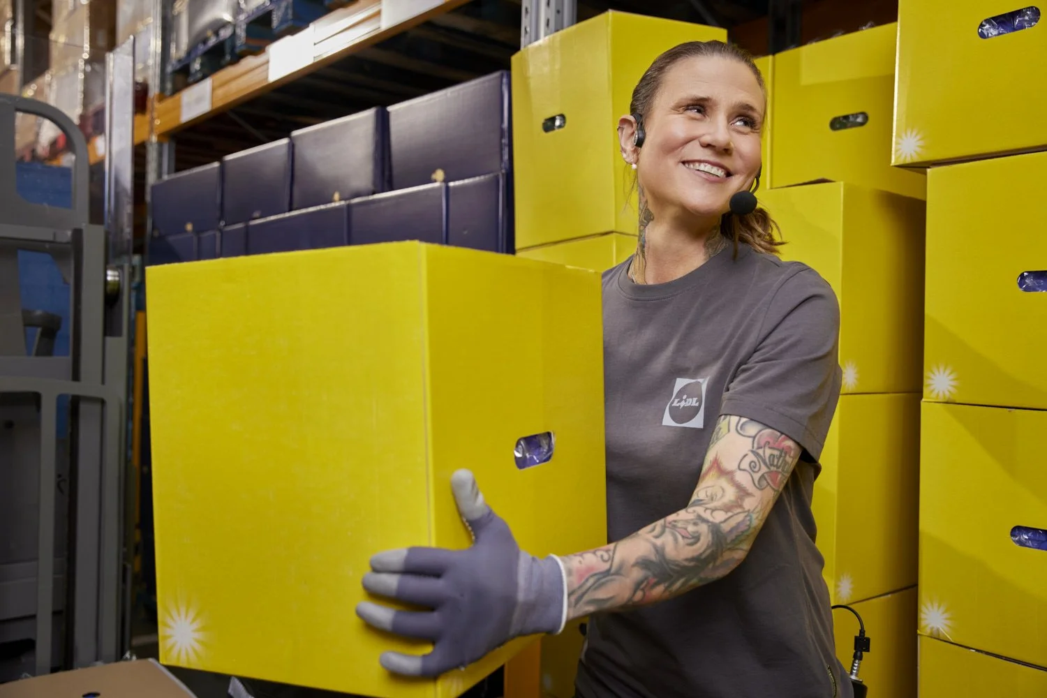 A woman with tattoos and a headset, wearing gray gloves, is smiling while handling a yellow box in a warehouse filled with stacked yellow boxes.