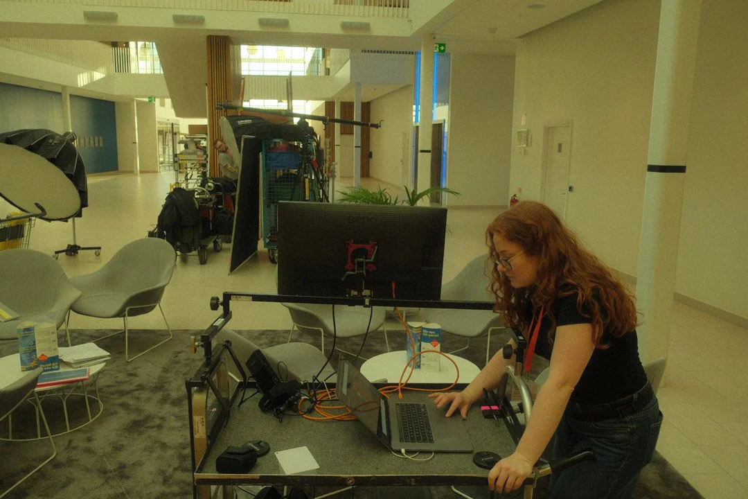A woman with curly red hair and glasses working at a table with a laptop and large monitor in a modern indoor space, likely a conference or event setup.