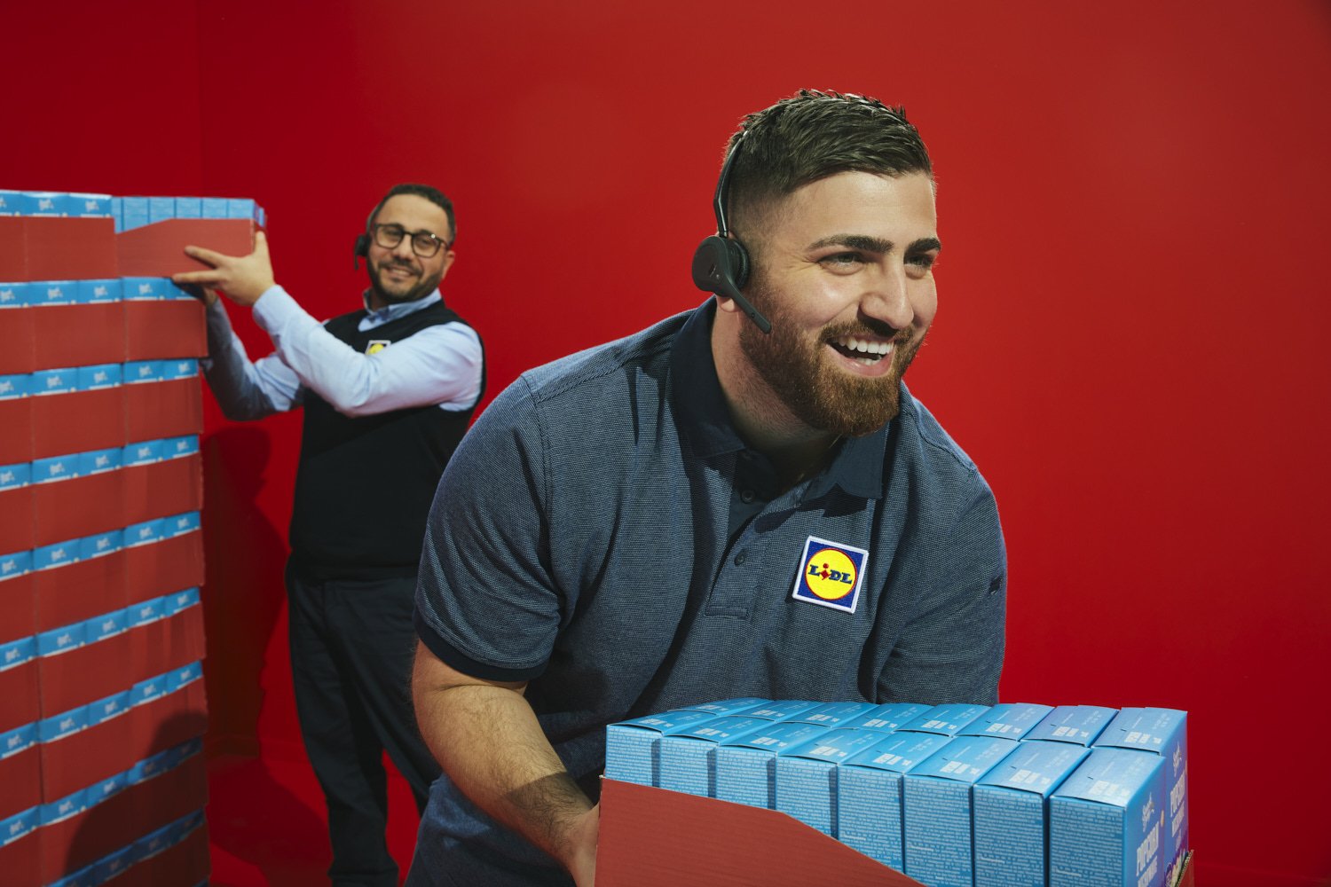 Two employees of Lidl store, a man with a beard and short dark hair, smiling and holding a large box of blue stacked product, and another man with glasses in the background, stacking red boxes against a red wall, wearing a headset and Lidl uniform.