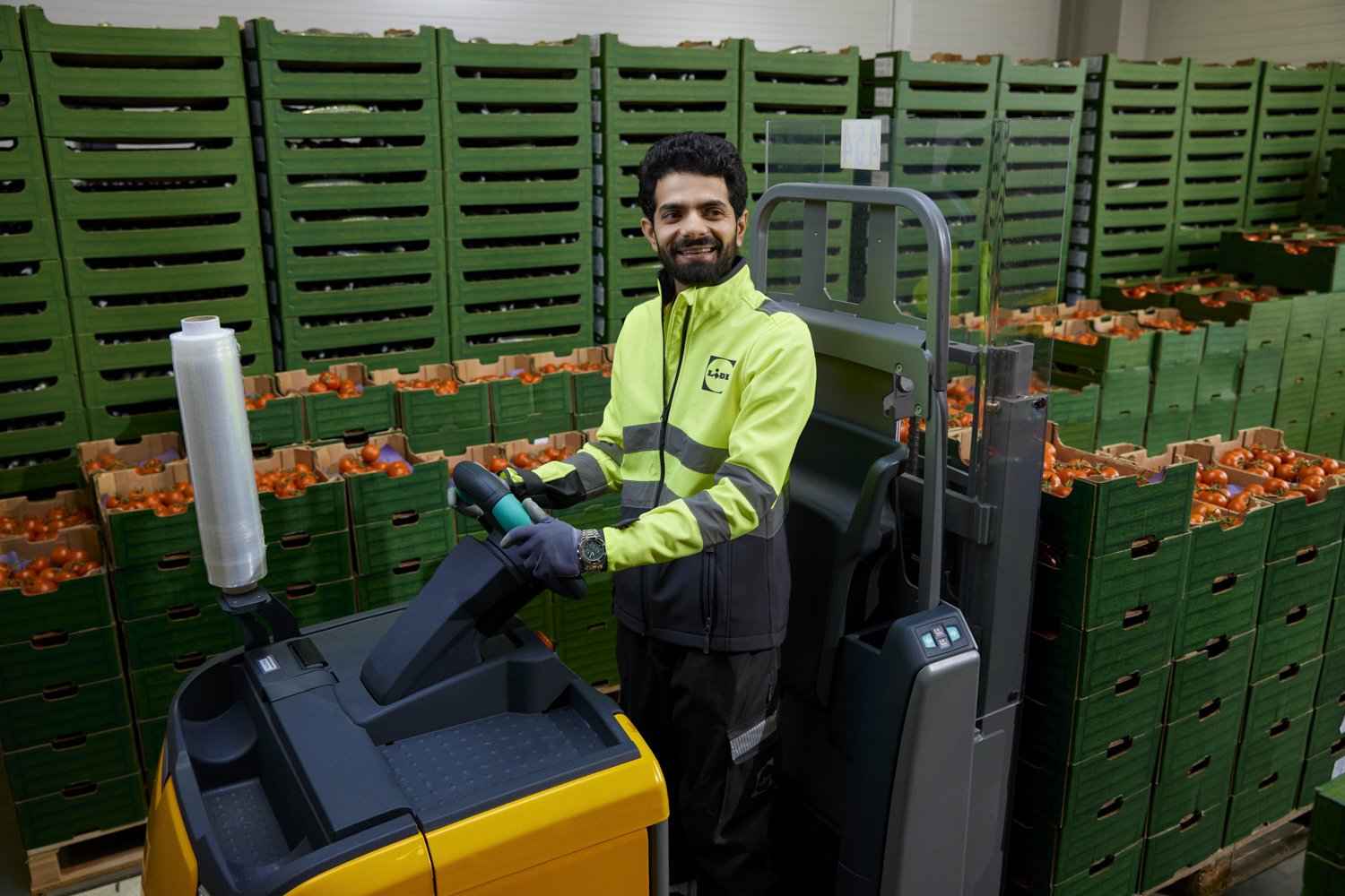 A man operating a yellow forklift in a warehouse surrounded by green crates of tomatoes.