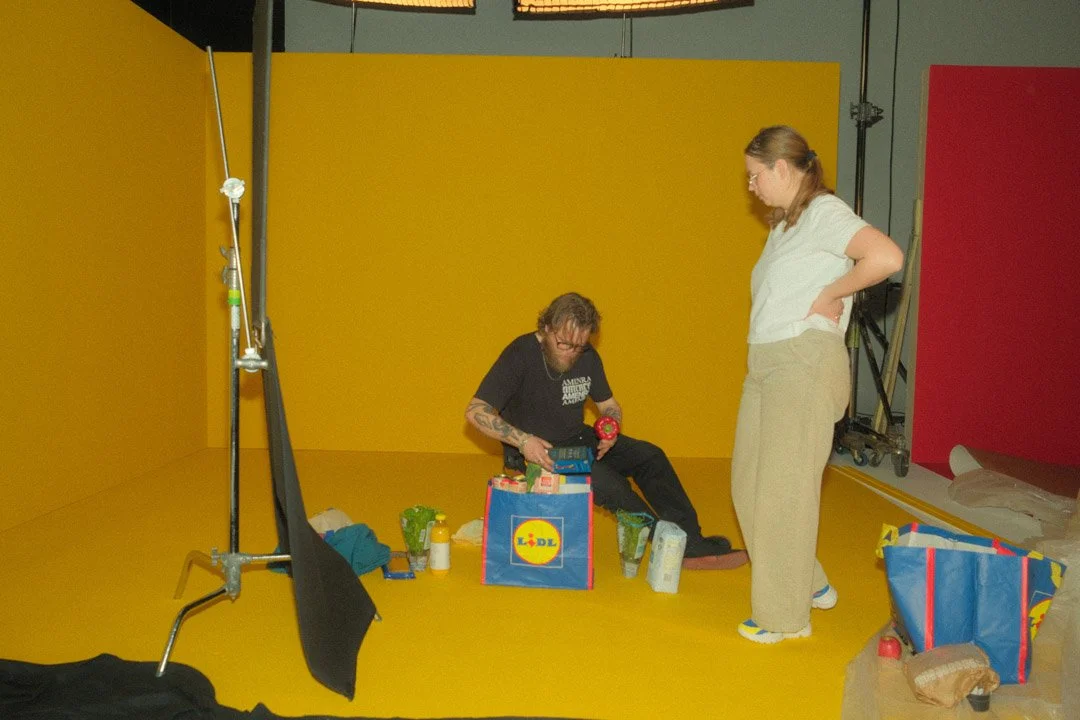 People preparing items in a yellow photo studio with red and yellow background panels, surrounded by bags, bottles, and equipment.