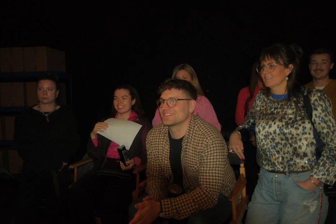 Group of people indoors with dark background, some sitting and some standing, engaged in conversation or listening.