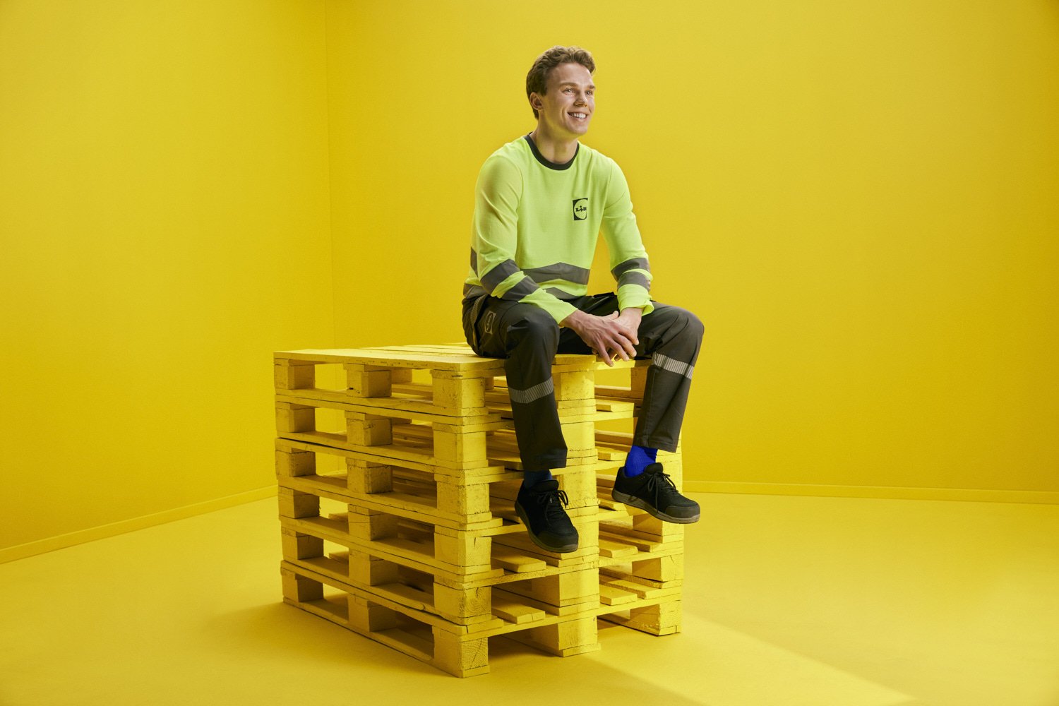 A young man in a high-visibility yellow safety jacket and black work pants sitting on top of yellow wooden pallets, smiling against a yellow background.