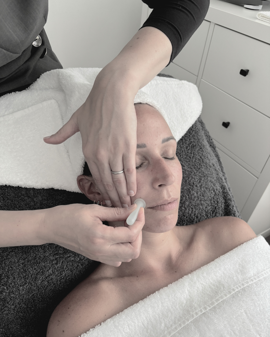 A woman with closed eyes receiving a facial treatment with a jade roller at a spa or clinic, lying on a treatment table with towels.