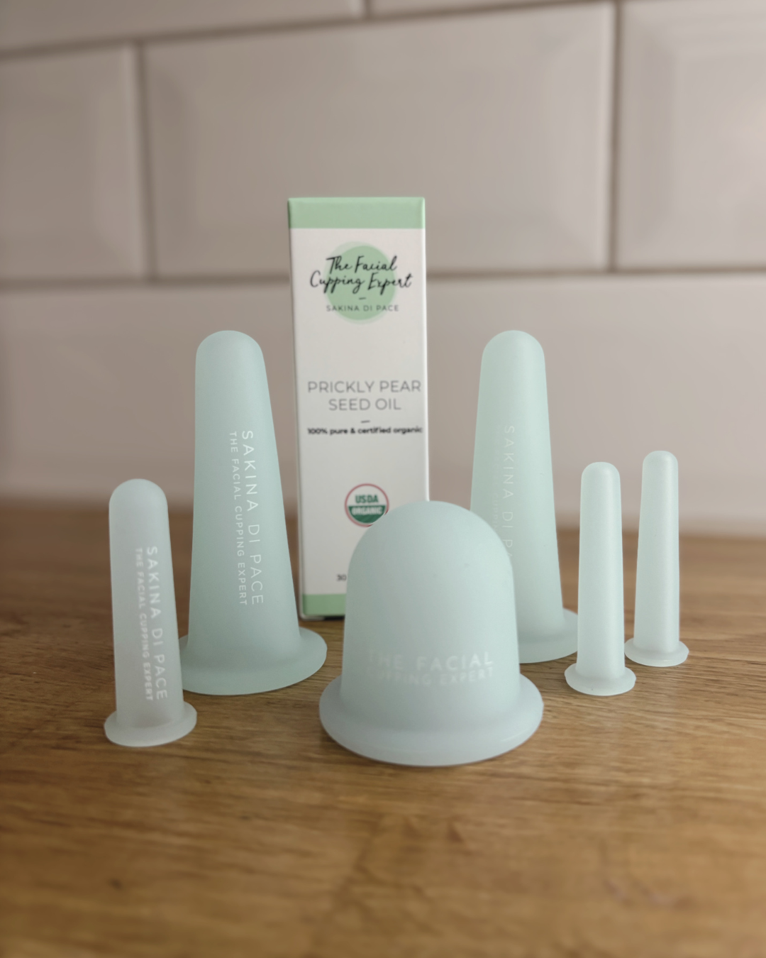 A collection of facial cupping tools and a box for prickly pear seed oil on a wooden surface, with a tiled wall in the background.