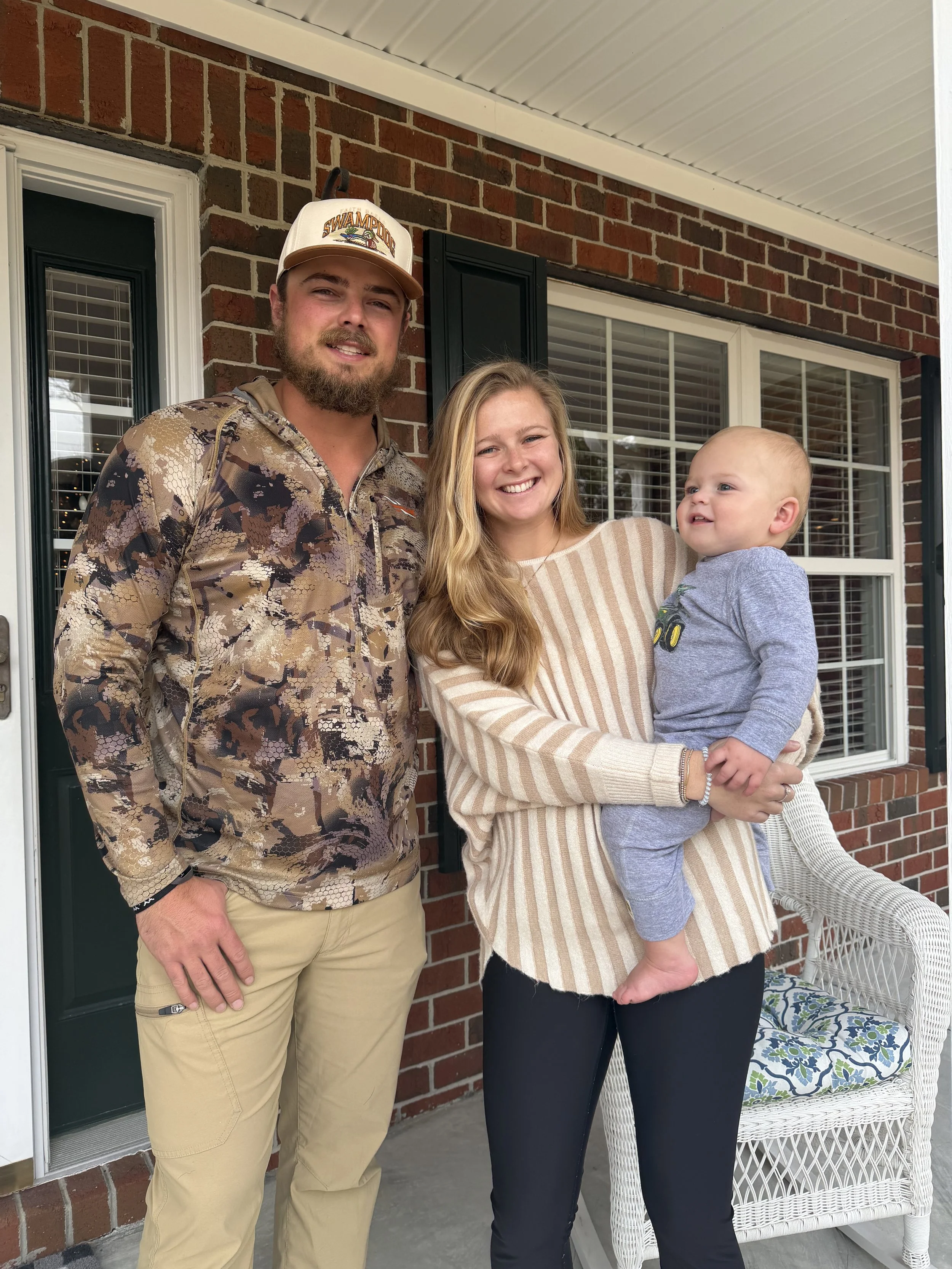 A woman and a man holding a newborn baby in a cozy, well-lit room with light blue walls. The woman is smiling down at the baby, who is dressed in light blue, while the man is looking lovingly at the baby and wearing a white cap and patterned shirt.