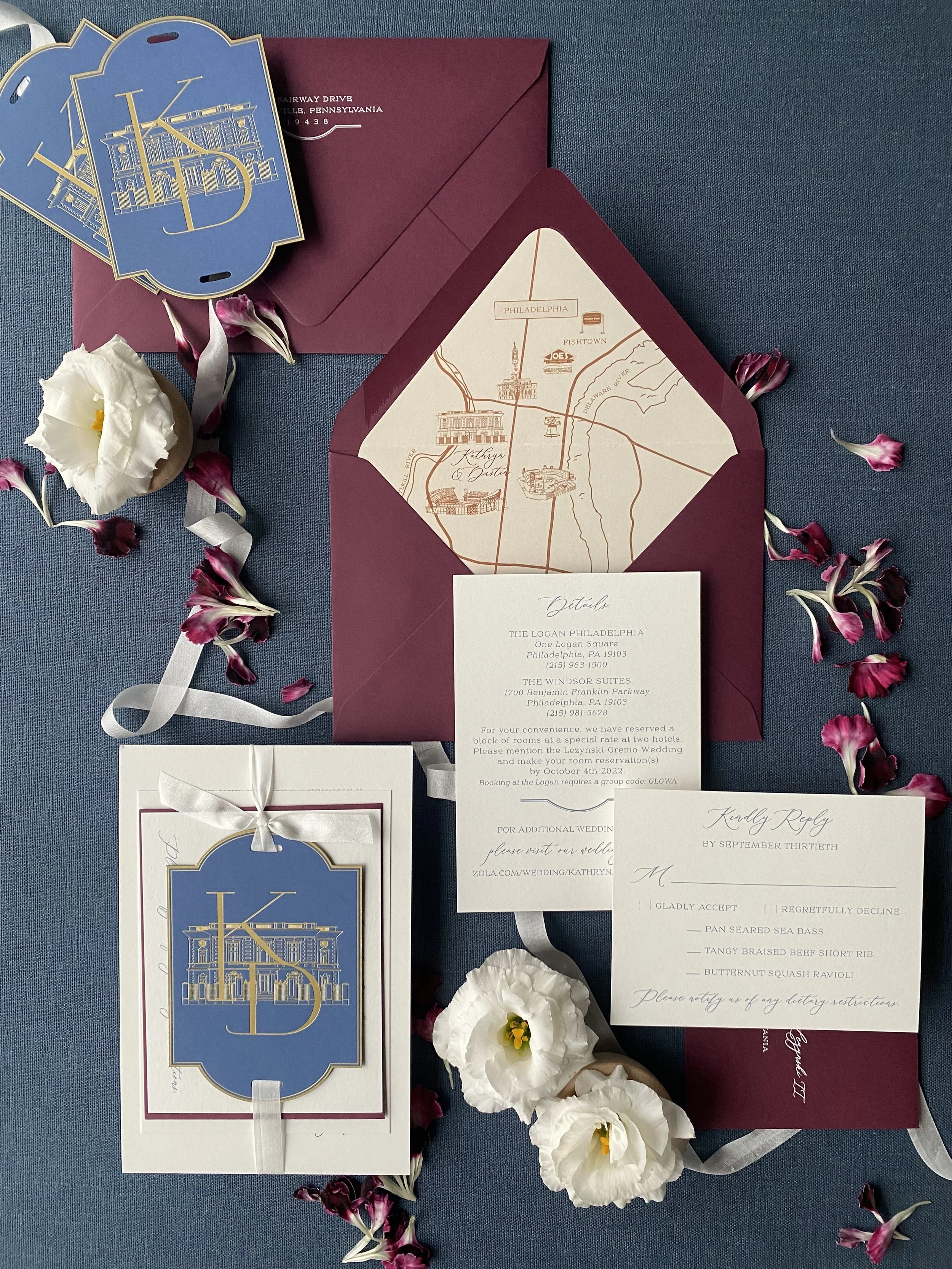 Burgundy and blue custom wedding invitation with a gold monogram atop a gold venue illustration of the College of Physicians in Philadelphia. The envelope liner is a map of the couple's favorite places in Philly. The suite is atop a blue background.