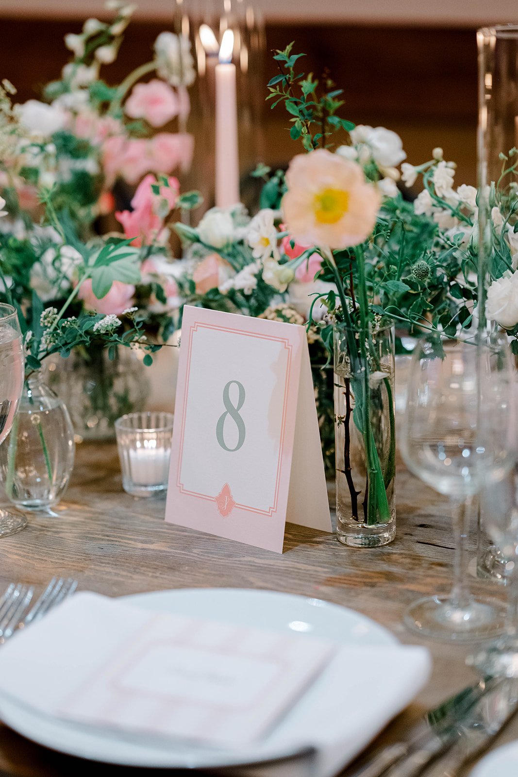 A wedding table number with a peach patterned frame and soft green text. Nestled among peach, pink, and white flowers and greenery. A white place setting with a menu is in the foreground.