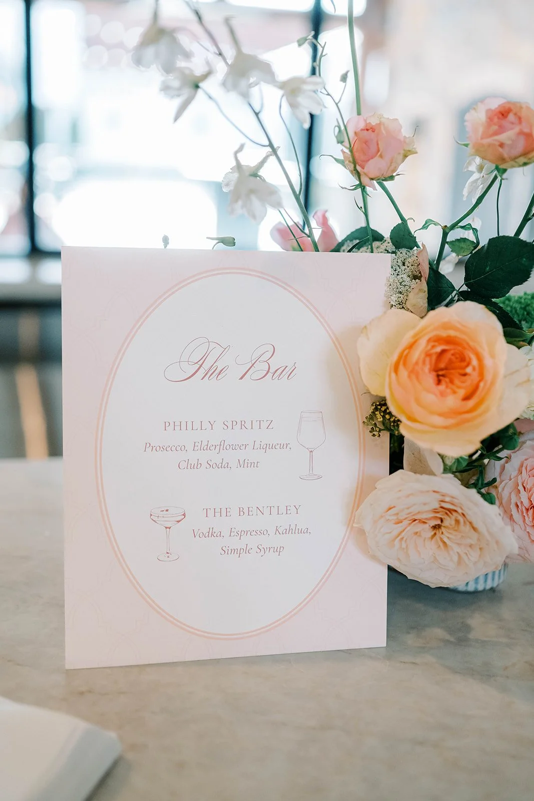 Custom bar signage for a spring wedding in peaches and pinks. Displayed on a bar next to a beautiful floral arrangement.