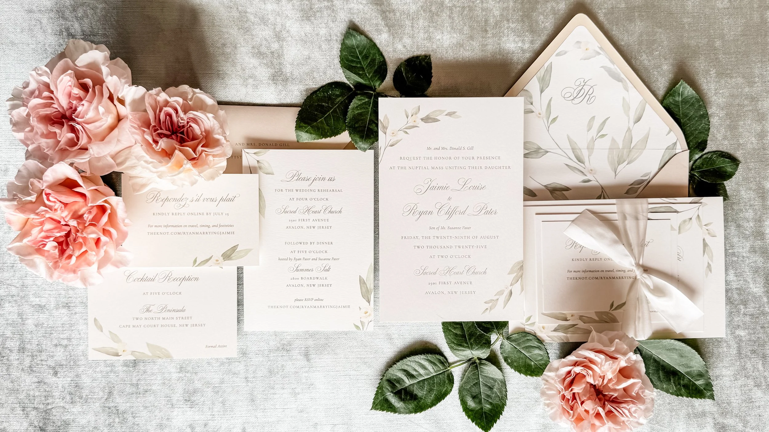 Flat lay of a wedding invitation suite with silk ribbon, watercolor greenery and beige envelopes on a velvet surface.  Surrounded by blush cottage roses and leaves.
