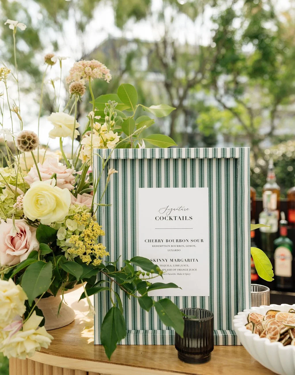 A beautiful fabric-covered custom drinks sign atop a wedding bar surround by flowers, garnishes, bottles and a candle.