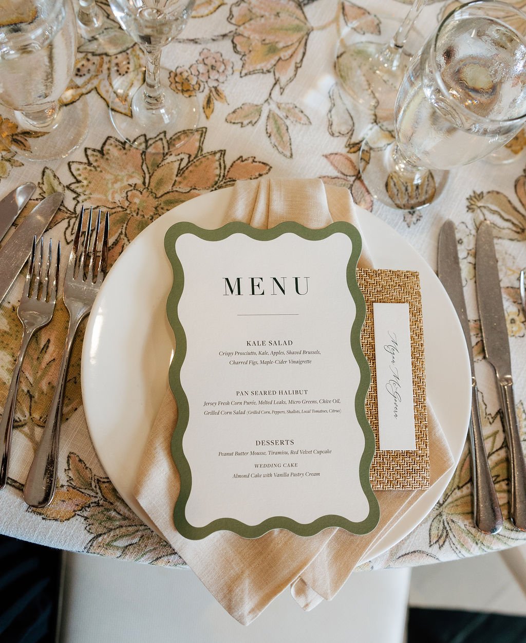 Beautiful table setting with pink and  green floral linens, and a place setting topped with a wavy edge menu with a green border and a rattan tag with guest names for a place card by Stone Hill Paperie
