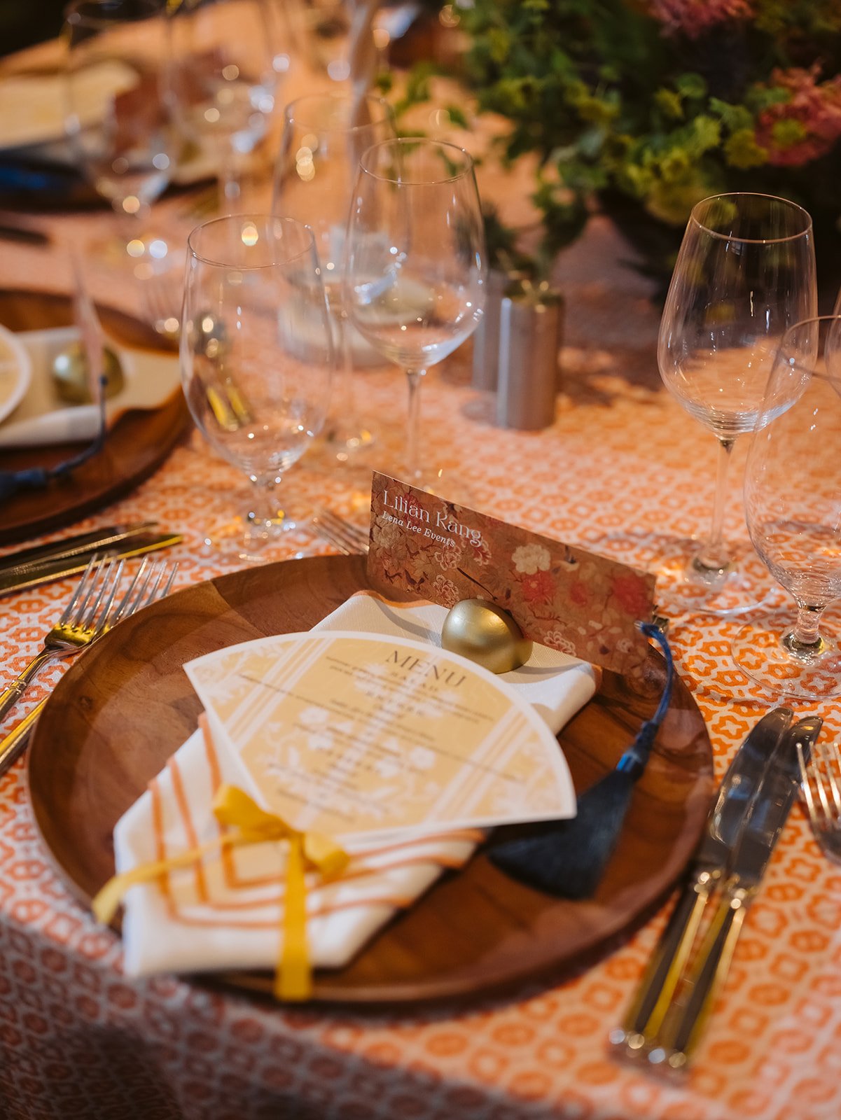 A die cut menu in the shape of a fan with a bow at the bottom and a subtle asian pattern in a soft yellow print on a plate topped with a napkin and a place card that doubles as a bookmark.