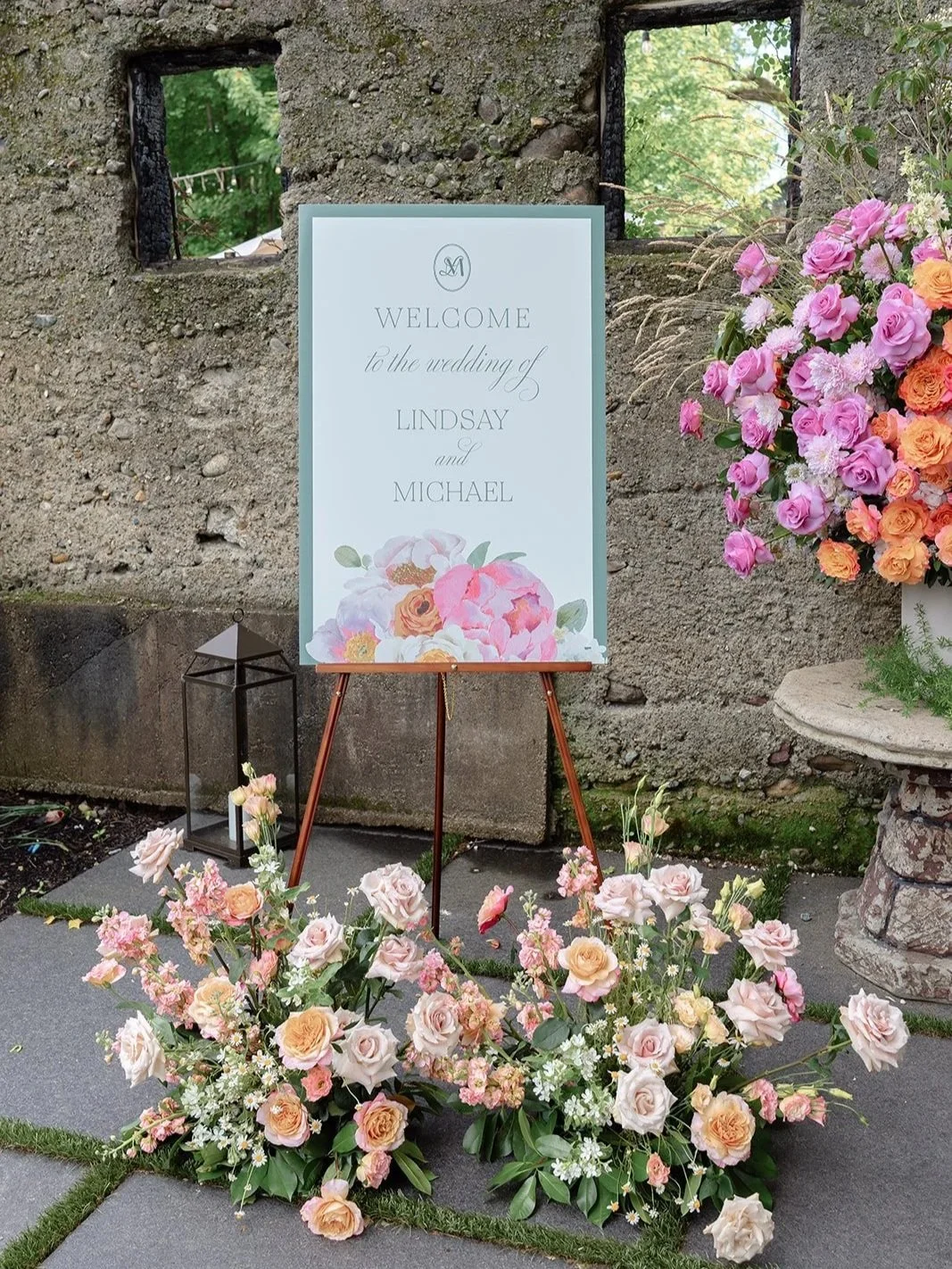 custom welcome sign with sage green text and watercolor flowers placed on an easel and surrounded by lush floral installations in vibrant colors. Placed at the entrance of Valley Rock Inn with rustic stone wall behind it.
