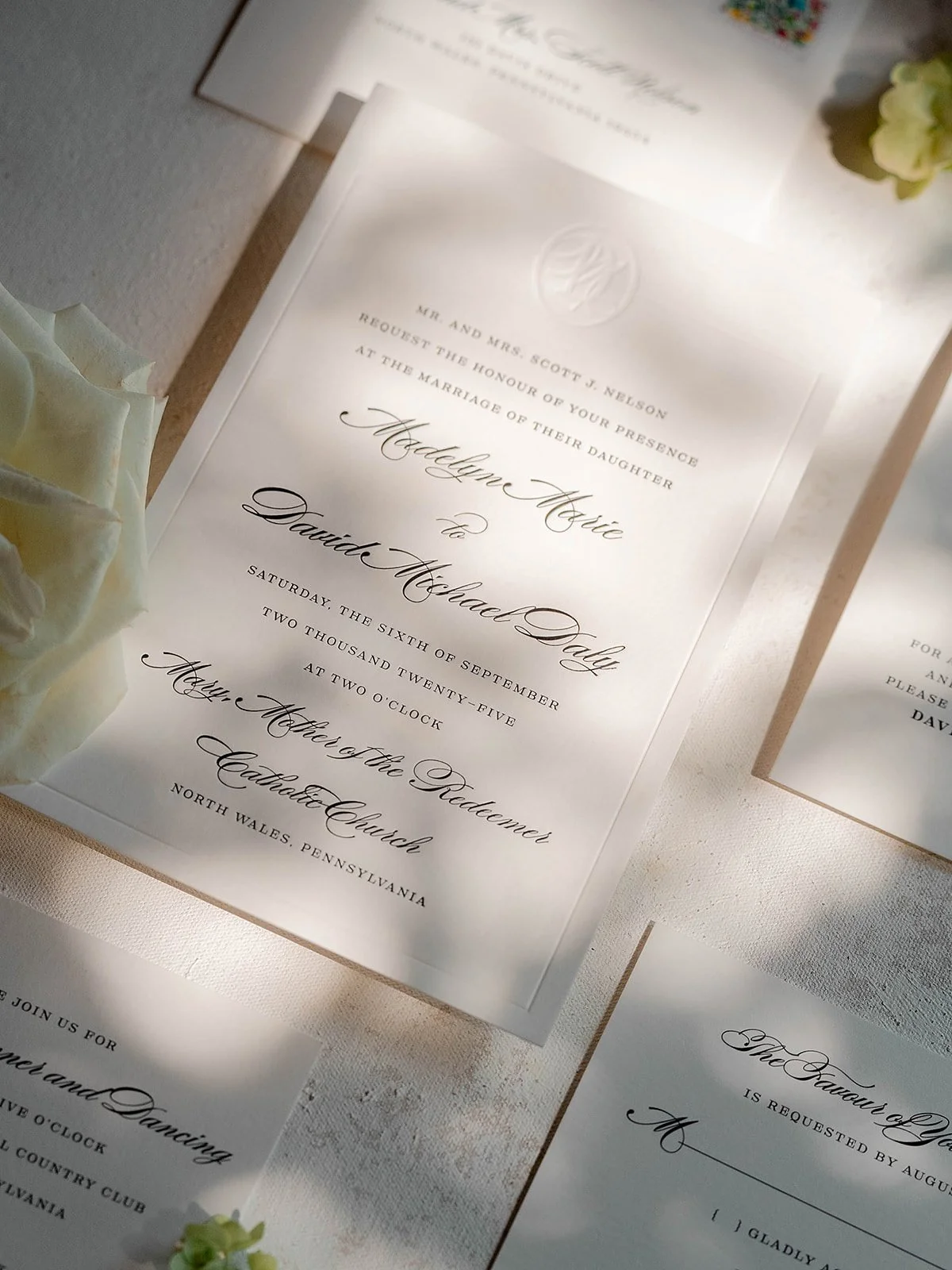 A wedding stationery with dappled light. The invitation is letterpress and debossed. There are a few flowers subtly entering the image from the edges.