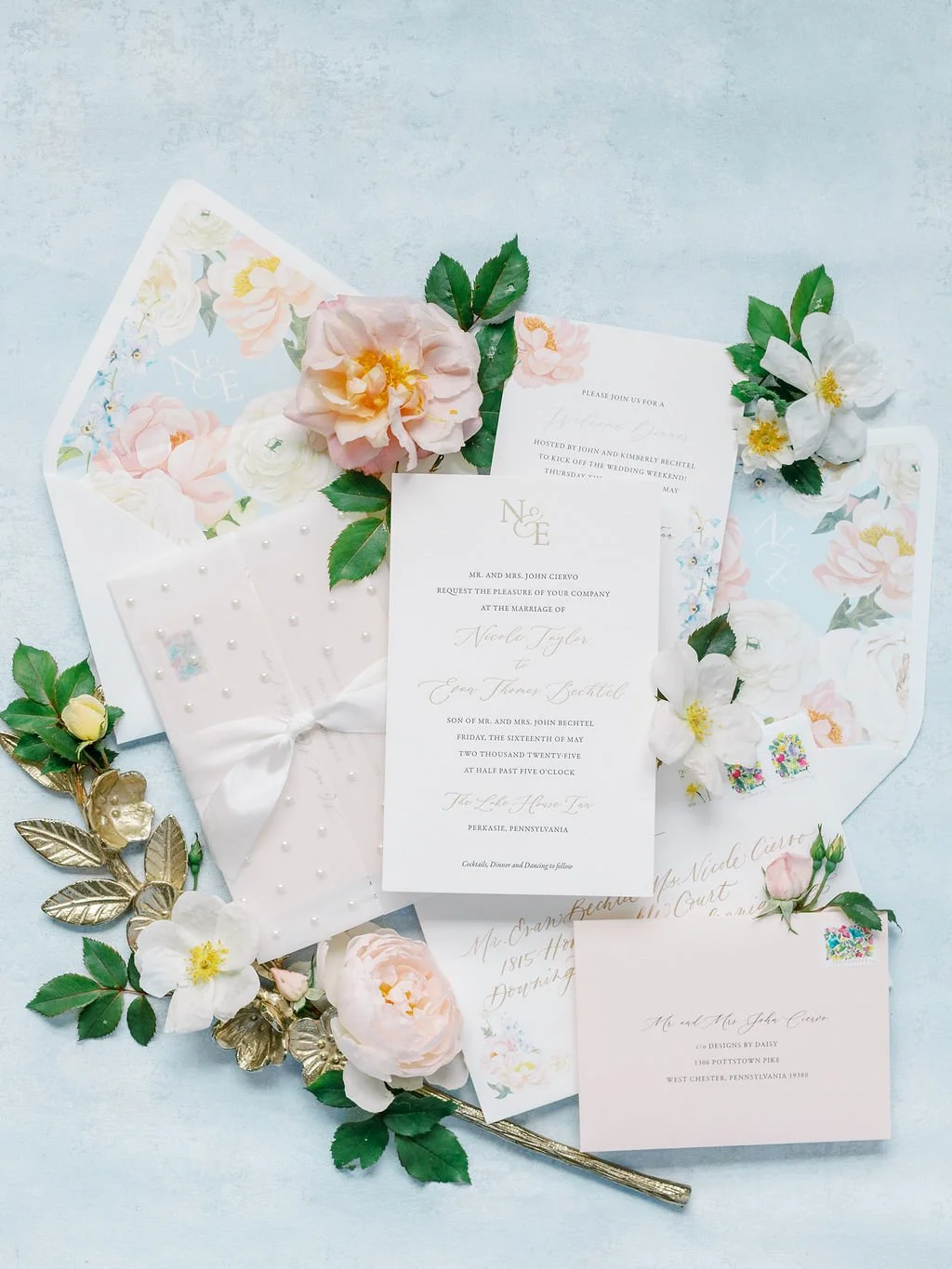 Foil and letterpress invitation with a pearl studded wrap, silk ribbon, and custom wax seal. The invitation is surrounded by gorgeous flowers and placed atop a soft blue background