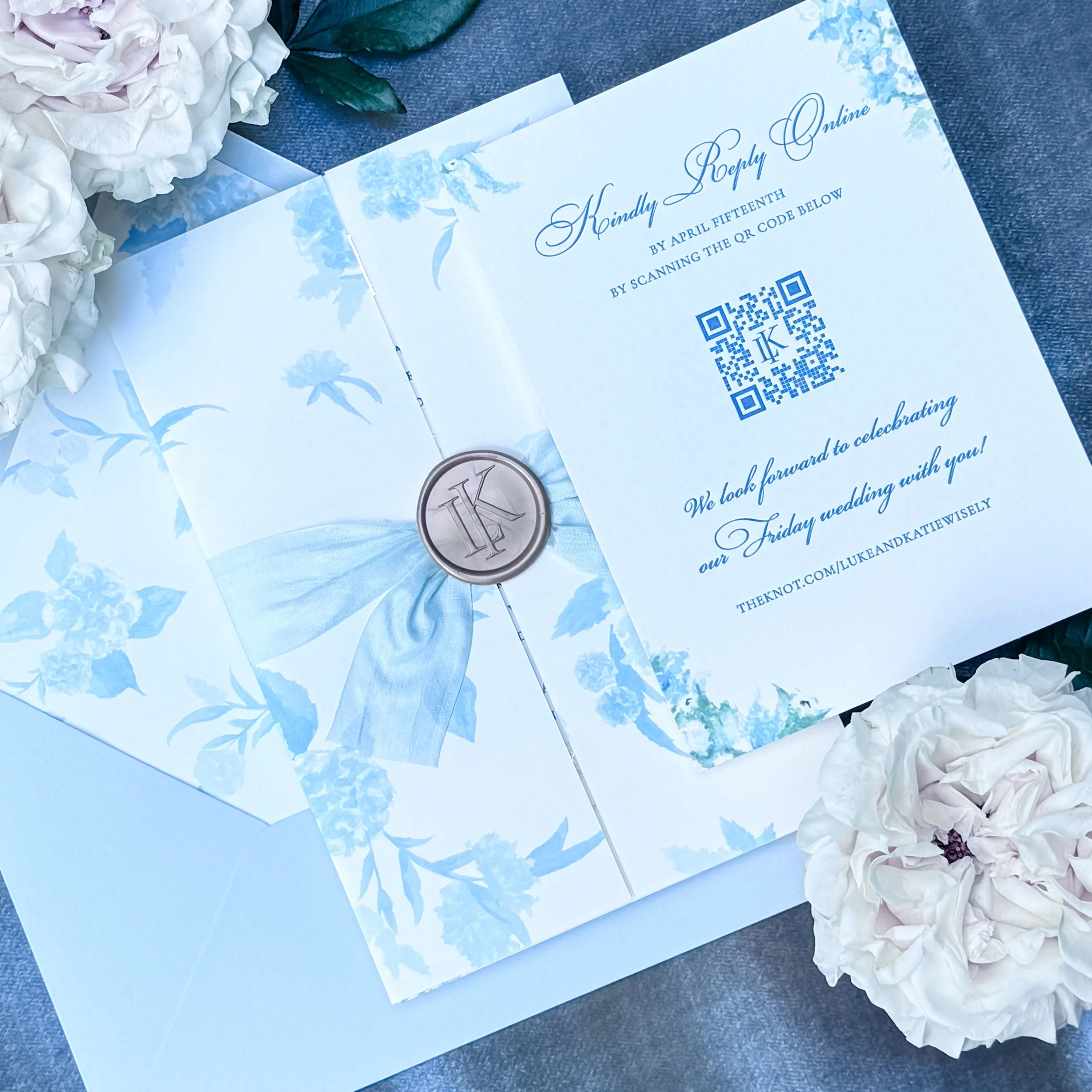 Wedding invitation with a light blue floral print, silk ribbon, and gold wax seal with the couple's monogram. Online response card with a QR code. Surrounded by white peonies on a blue background.