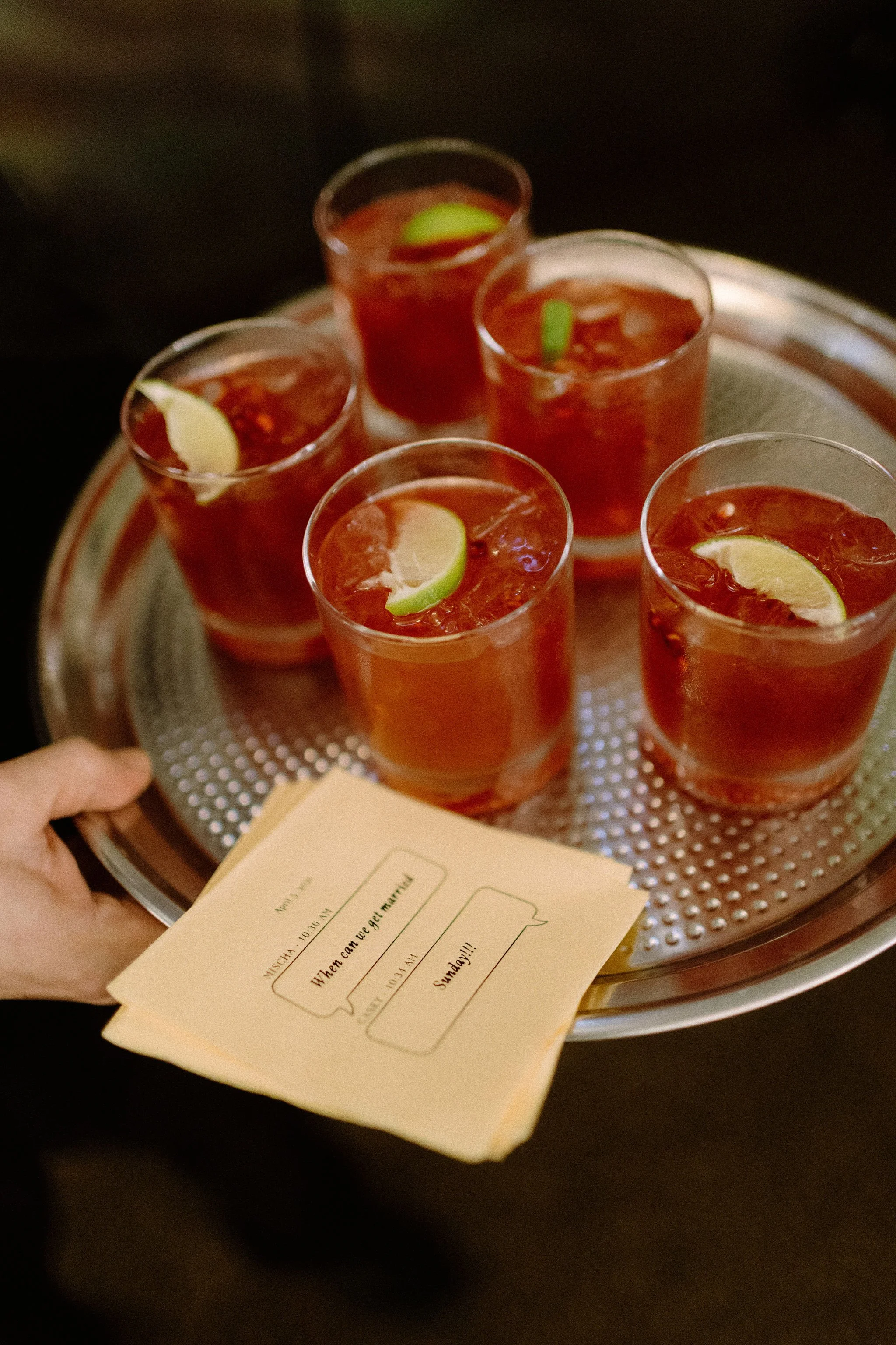 a hand holding a silver tray with 5 red cocktails with lime garnishes. Custom yellow cocktail napkins with actual text messages between a bride and groom while they were dating.