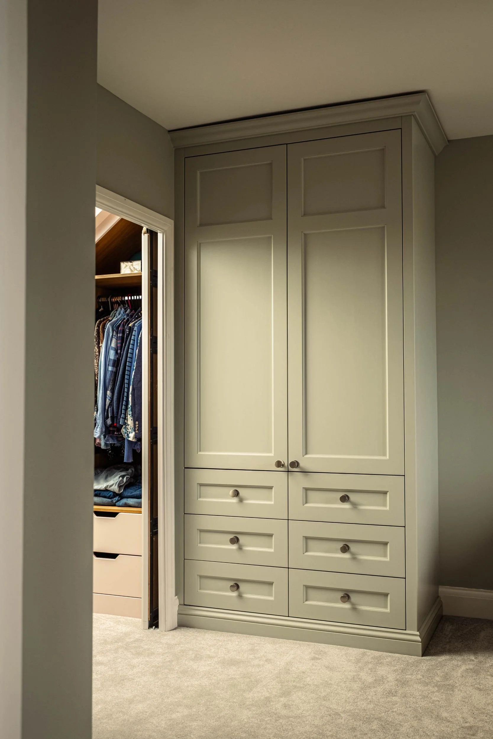 A made to measure fitted wardrobe with two Shaker style doors and six Shaker style doors. The wardrobe is painted green and to the left there is a walk in wardrobe with open shelving and hanging rail space and drawers.