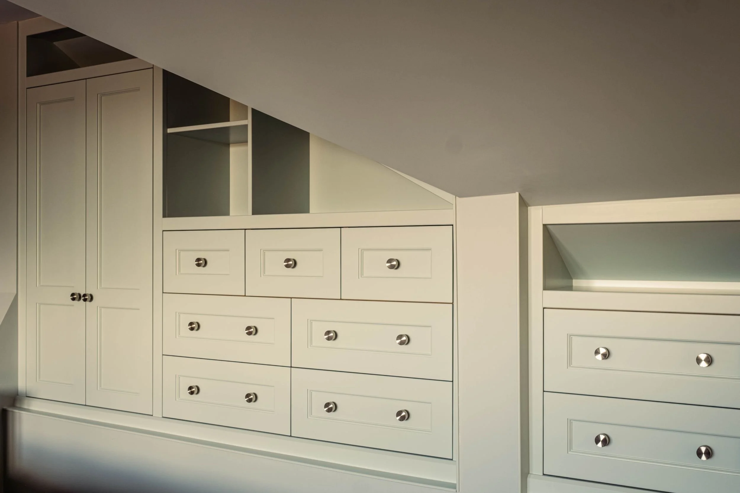A large made to measure fitted storage unit with a wardrobe, open shelving and drawer units, all built into a loft space with sloping ceilings.
