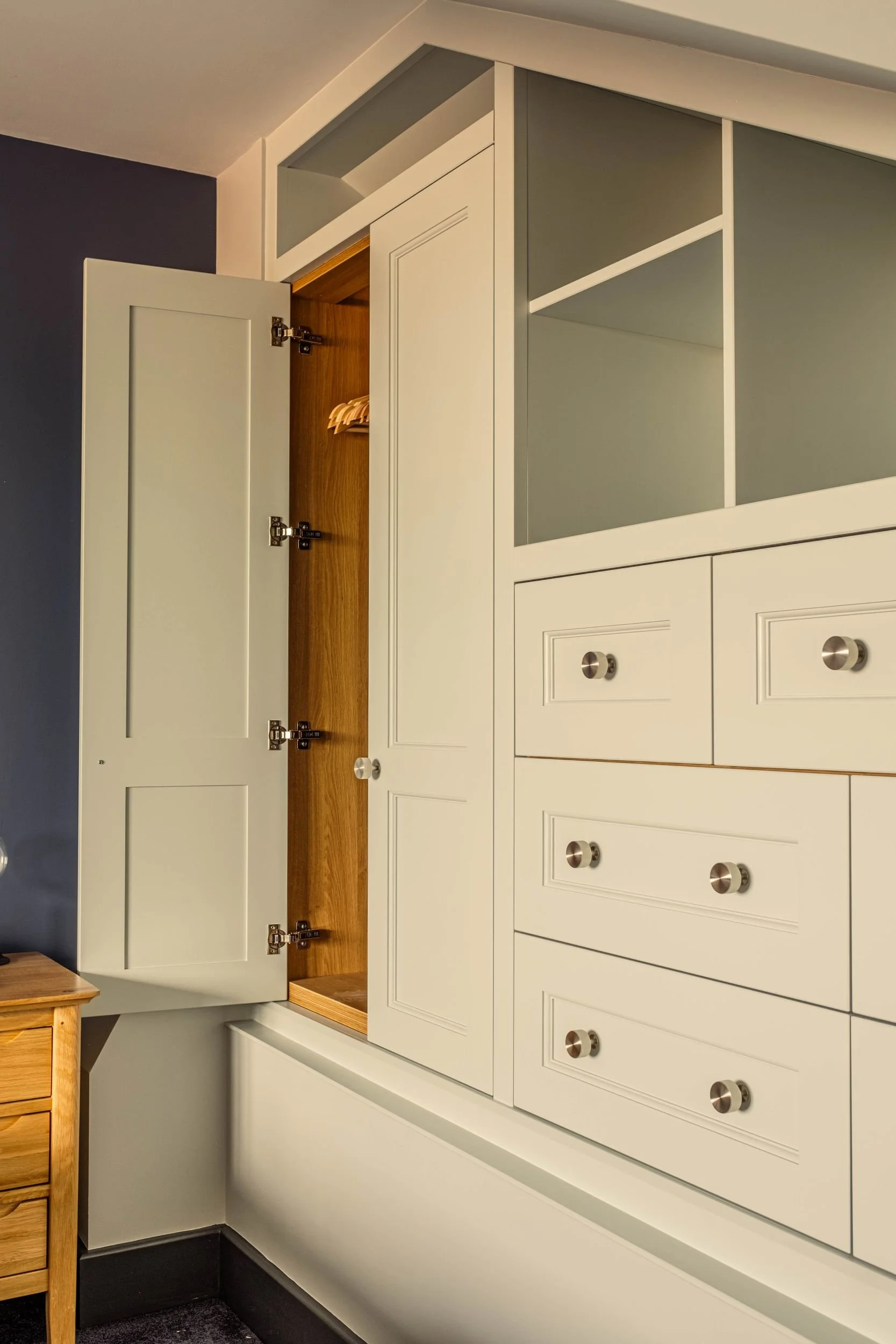 Open white built-in wardrobe with wooden interior and hanging space, adjacent to drawers and cabinets with silver knobs, in a modern room.