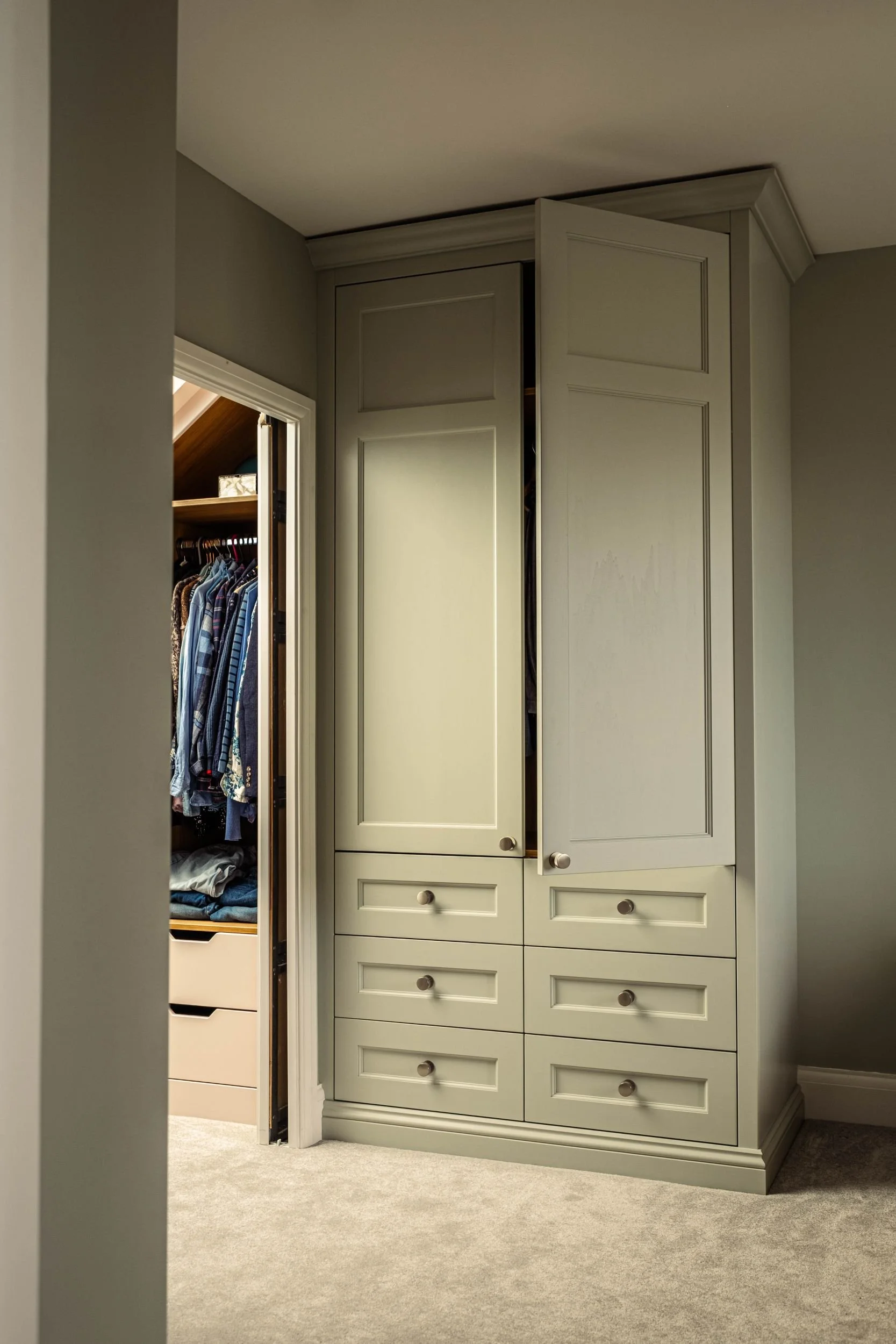A made to measure fitted wardrobe with two Shaker style doors and six Shaker style doors. The wardrobe is painted green and to the left there is a walk in wardrobe with open shelving and hanging rail space and drawers.