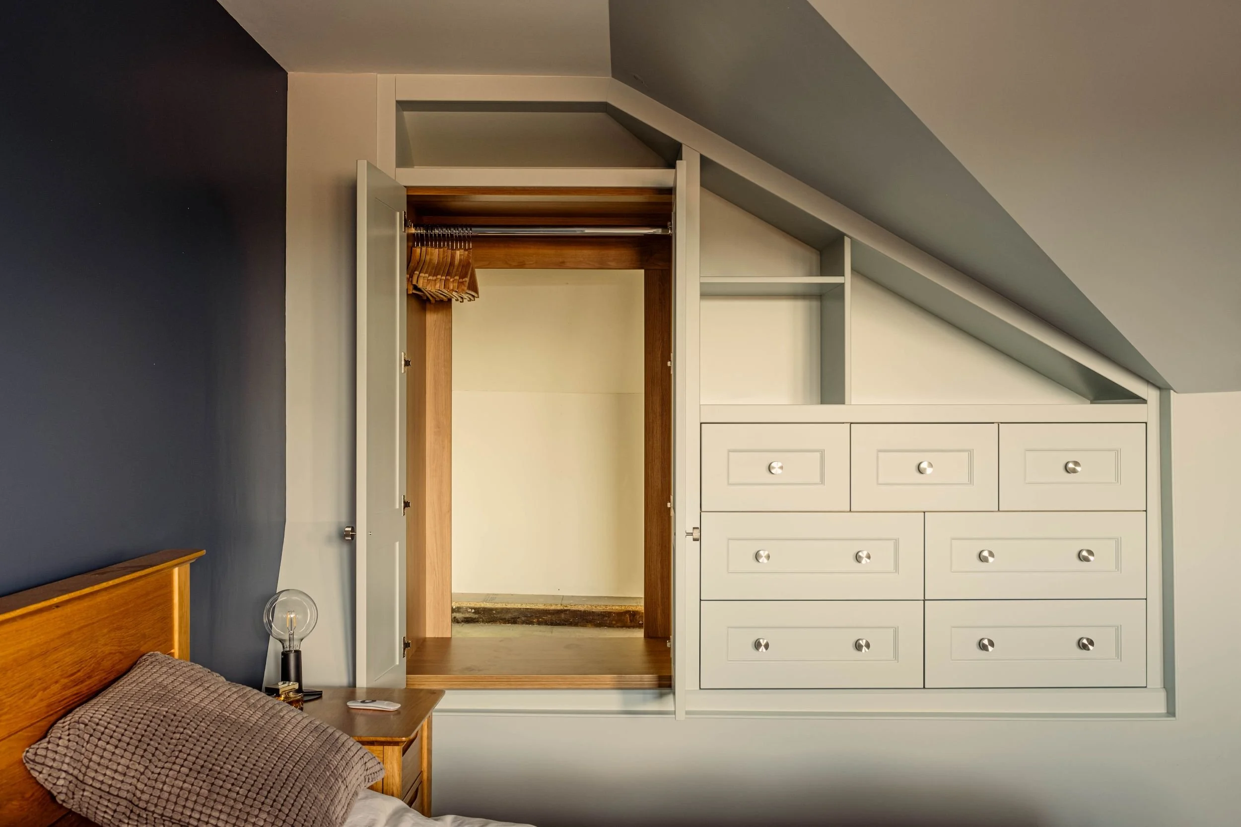 A made to measure fitted storage unit with a wardrobe, open shelving and drawer units, all built into a loft space with sloping ceilings. The wardrobe doors are open. Its a Shaker style design.