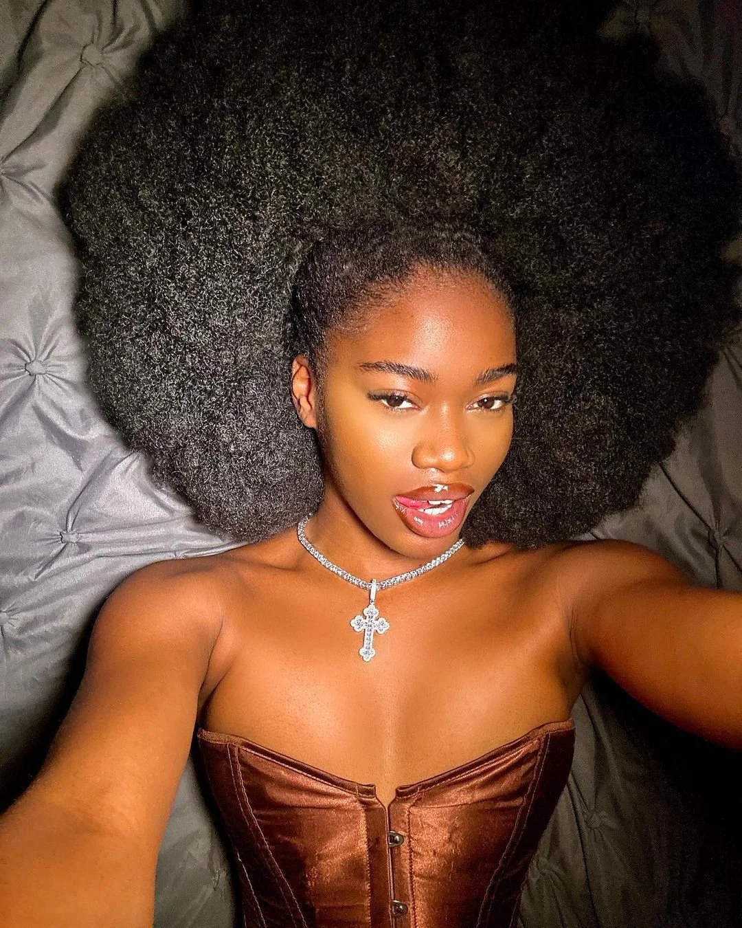 A woman with a voluminous afro hairstyle lying on a tufted gray surface, wearing a brown corset top and a silver cross necklace.