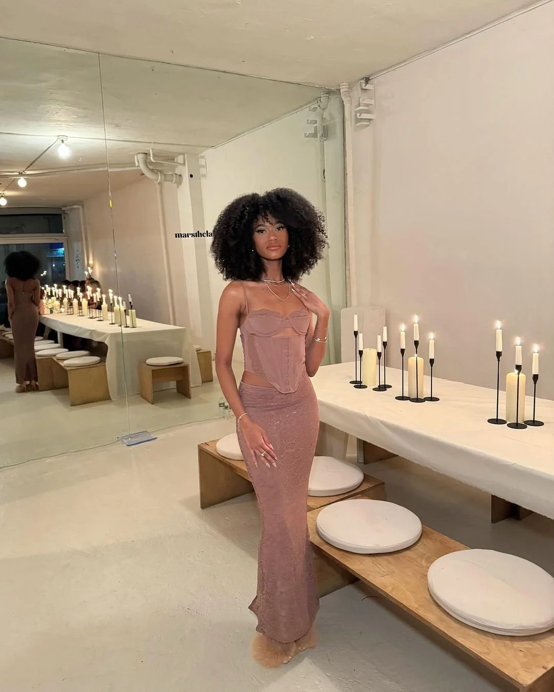 A woman with curly black hair and a pink dress standing next to a table with tall candles in a modern, minimalistic room with a mirror.