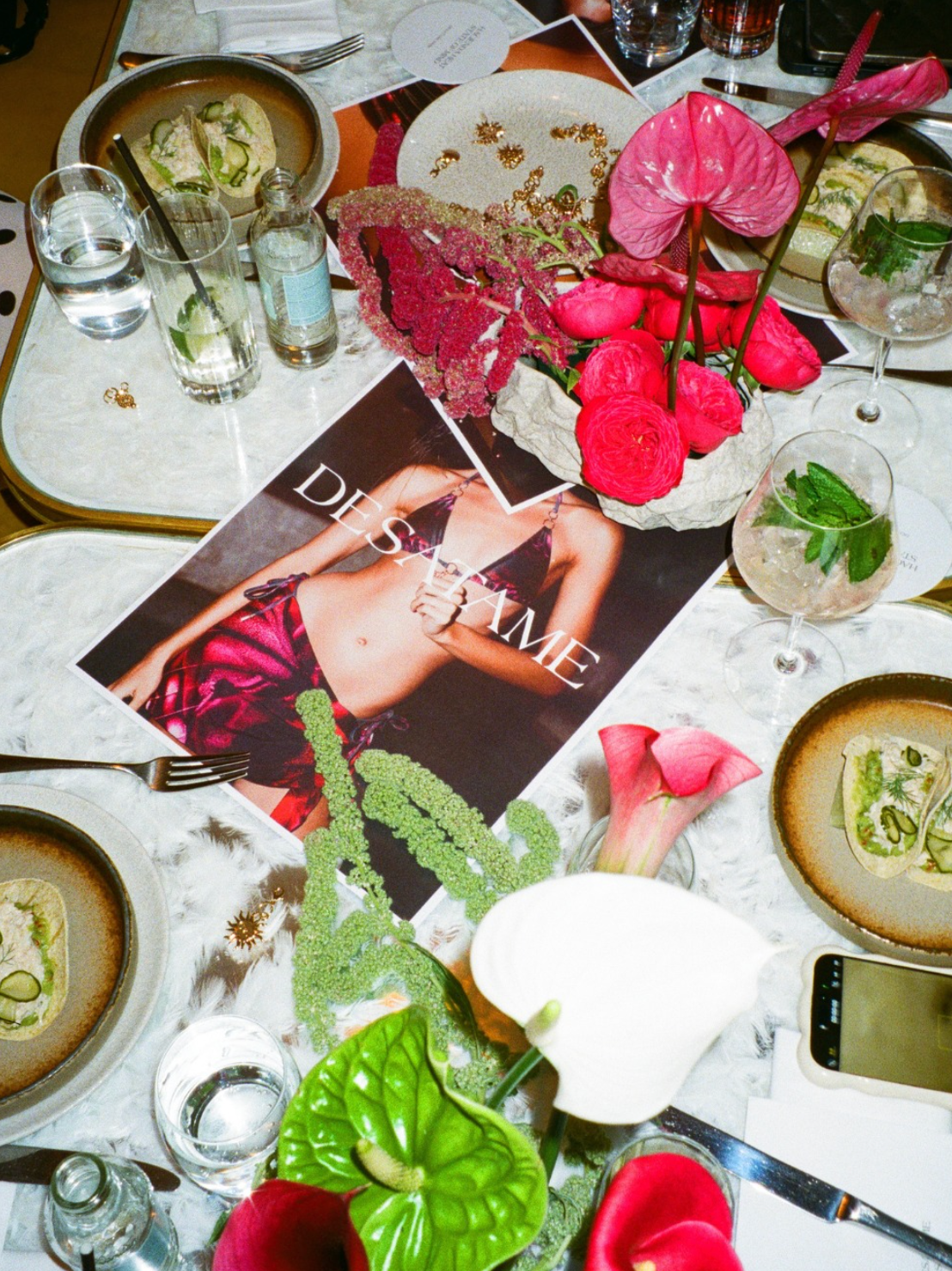 Table setting with floral centerpiece, plates of food, drinks, and a fashion magazine titled "DESI AME" featuring a woman in a pink outfit.