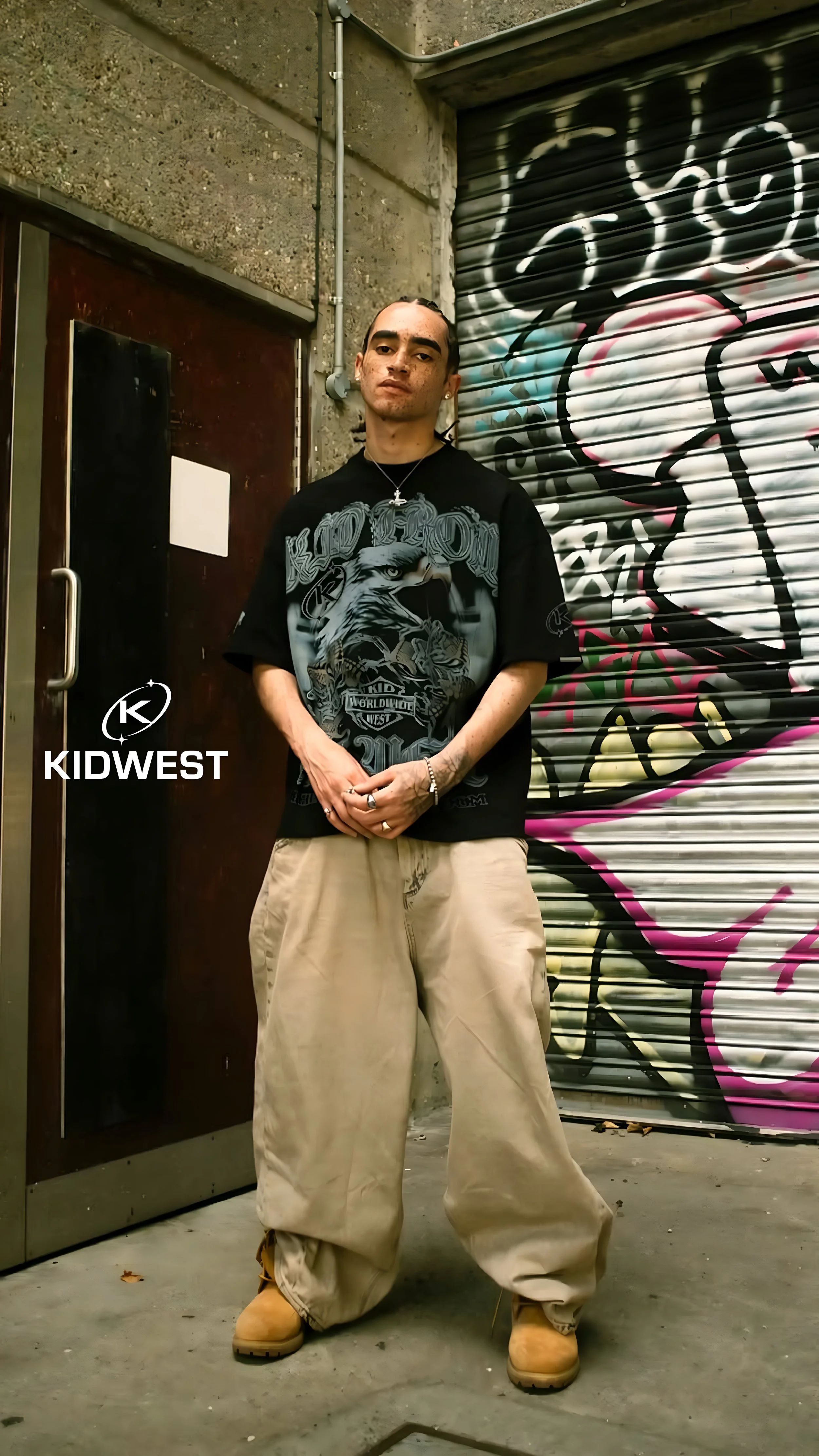 Young man with dark hair, tattoos, and a cross necklace standing in front of a graffitied metal shutter and concrete wall, wearing a black graphic t-shirt, baggy beige pants, and tan work boots.