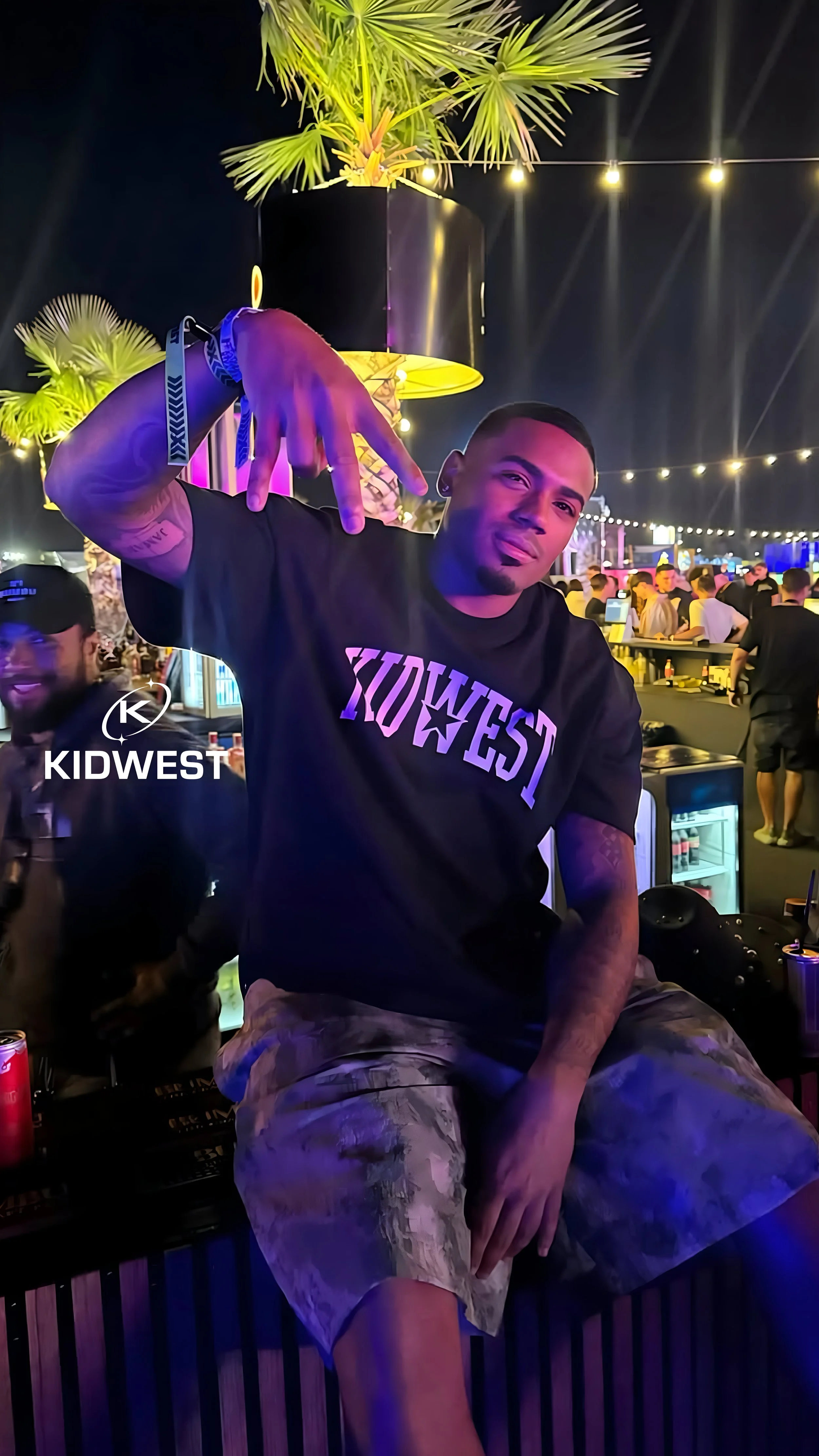 A young man making a peace sign with his right hand at a night outdoor party or bar with bright string lights, palm plants, and a crowd in the background.