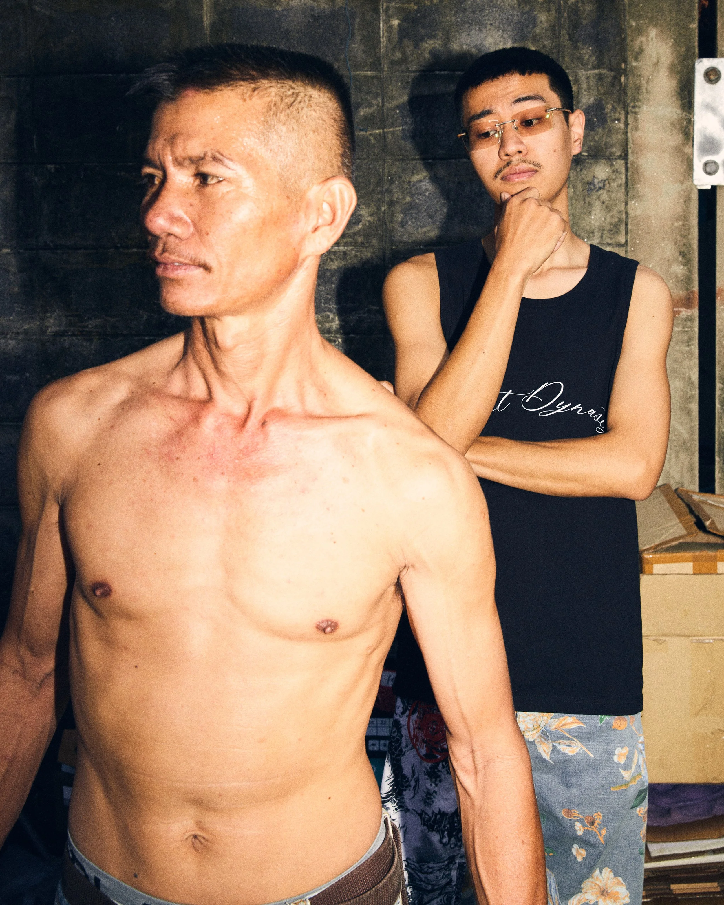 A shirtless man with short hair and a man in a black tank top with glasses standing behind him, both in an industrial setting with dark wall background.