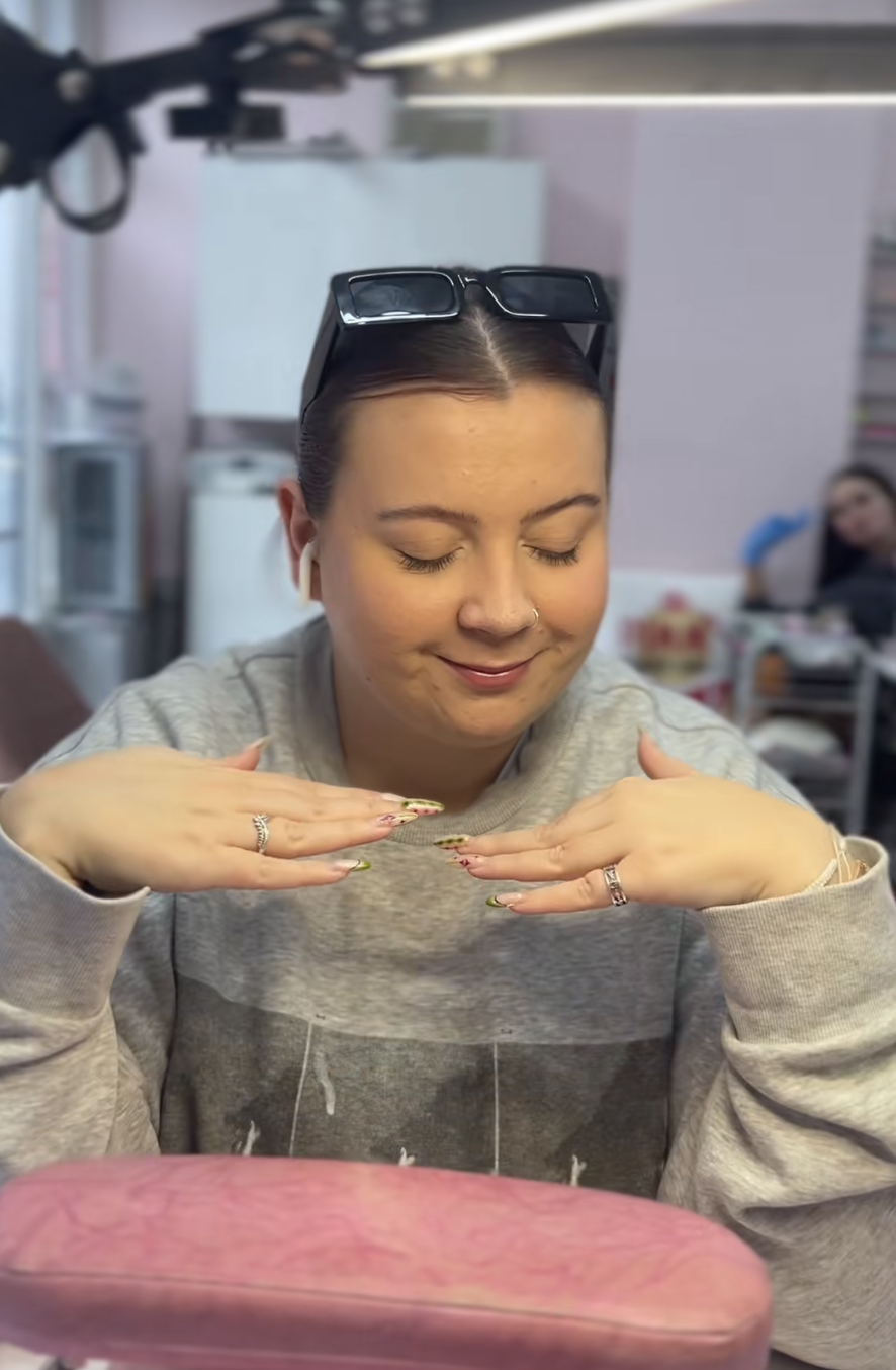 A woman with glasses on her head, closed eyes, and a nose ring, smiling and posing with her hands under her chin, wearing a gray sweatshirt. She is in a room with another person blurred in the background, sitting at a table.