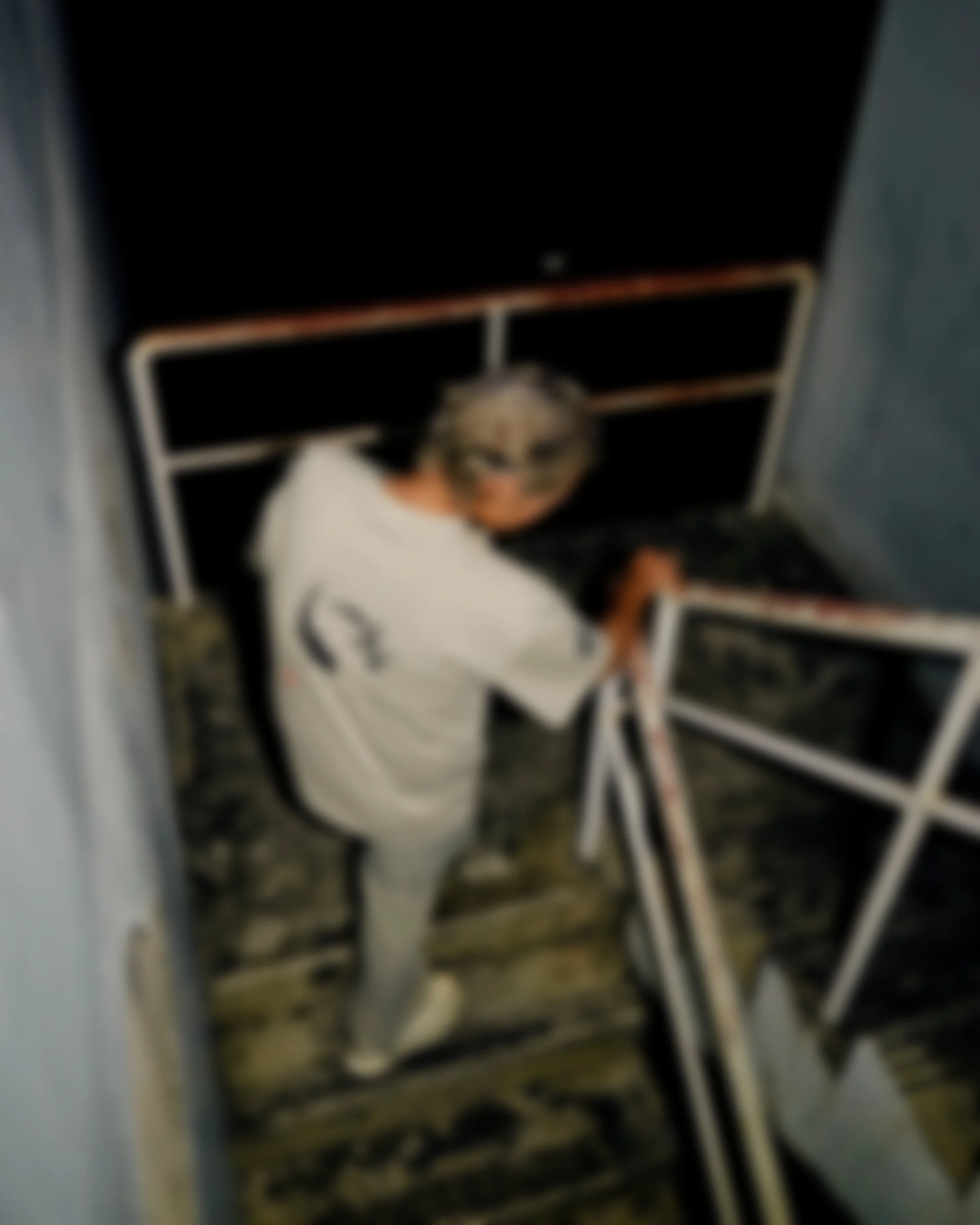 Person with short gray hair wearing glasses and a white shirt standing on a wooden staircase, looking up at the camera, next to a railing and a black chalkboard or sign.