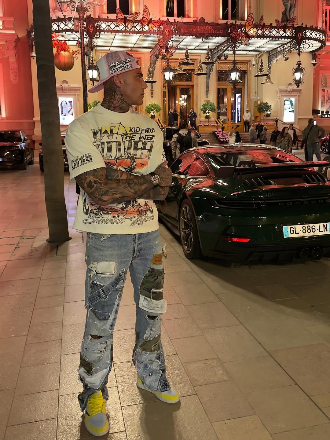 A tattooed man wearing a white graphic T-shirt, distressed patchwork jeans, yellow sneakers, a white cap, and a watch stands in front of a black sports car outside a brightly lit hotel or casino entrance decorated with butterflies and lights. People are walking and standing nearby.