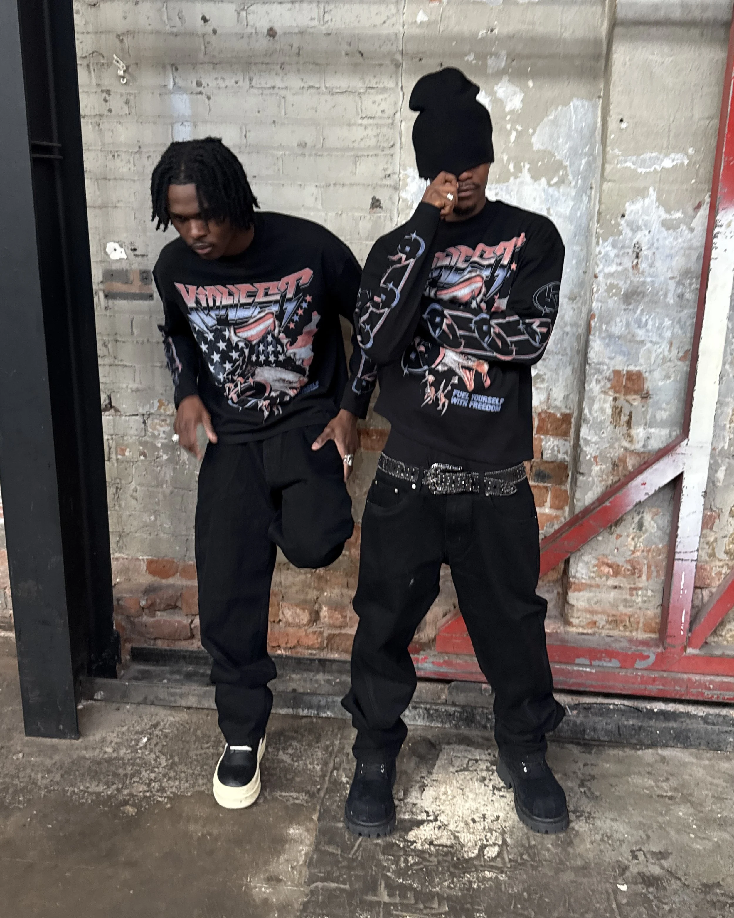 Two young men standing against a damaged brick wall, both wearing black graphic long-sleeve shirts and black pants. The man on the left has dreadlocks and white shoes, while the man on the right has his face partially covered with a beanie and is wearing black boots.