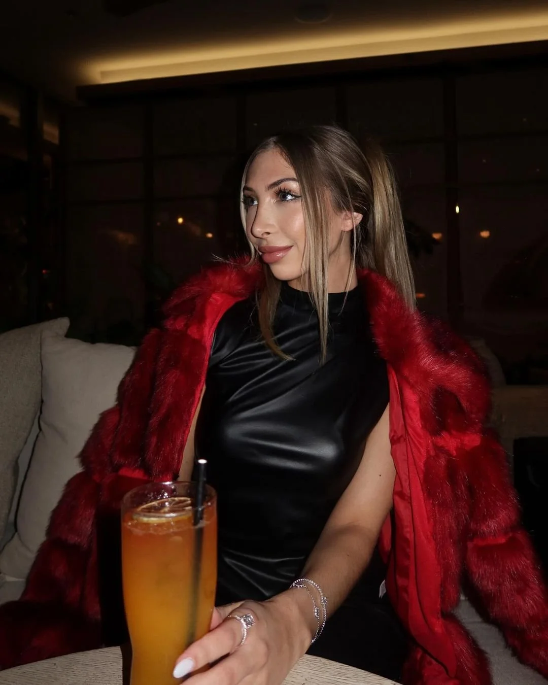 A young woman wearing a red fur coat and black satin dress sitting at a table with a tall orange cocktail drink in a glass with a black straw, in a dimly lit indoor setting.