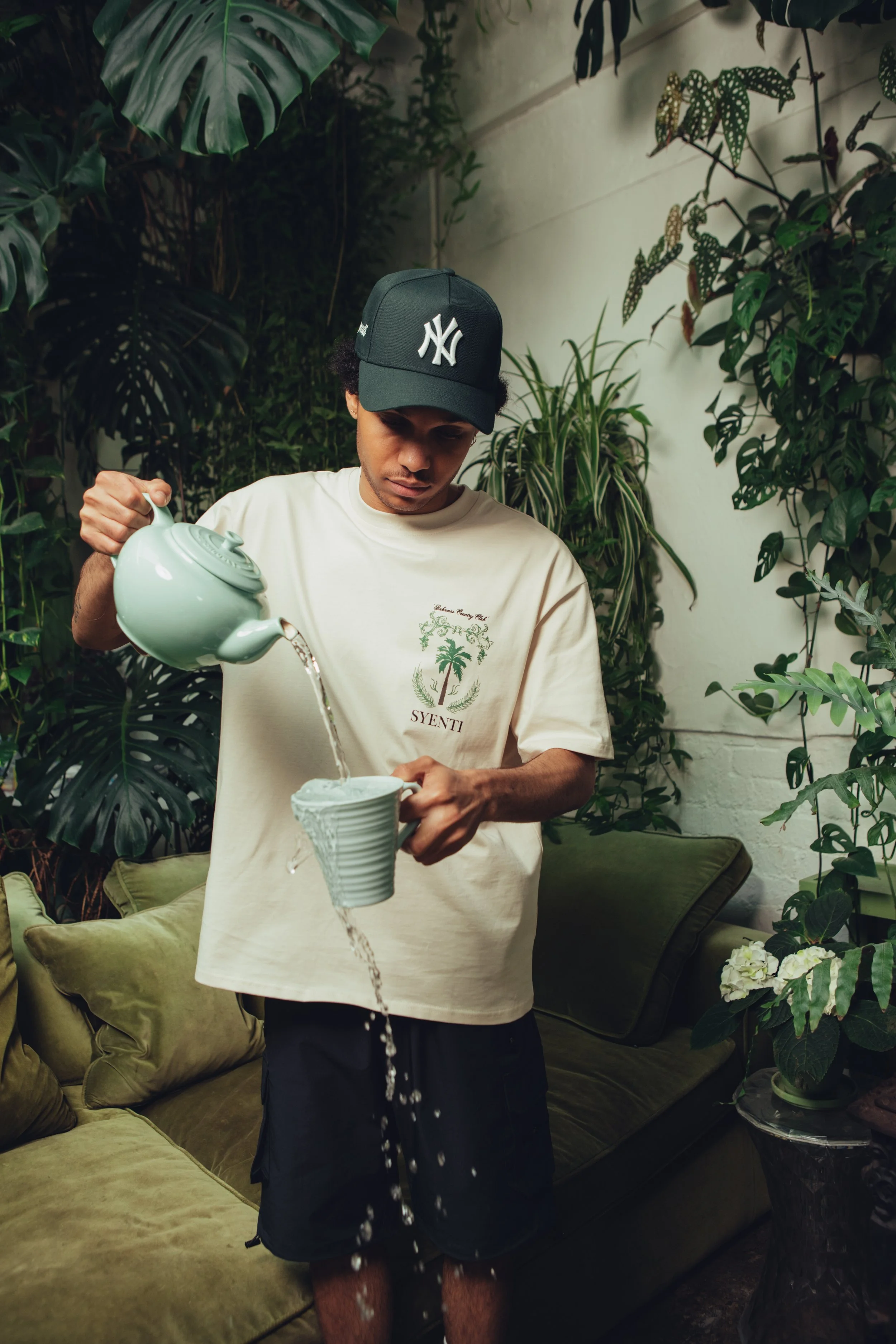 A man wearing a black New York Yankees cap and a beige t-shirt is pouring water from a mint green teapot into a striped mug. He is standing inside a room with lush green plants and a green sofa with matching pillows.