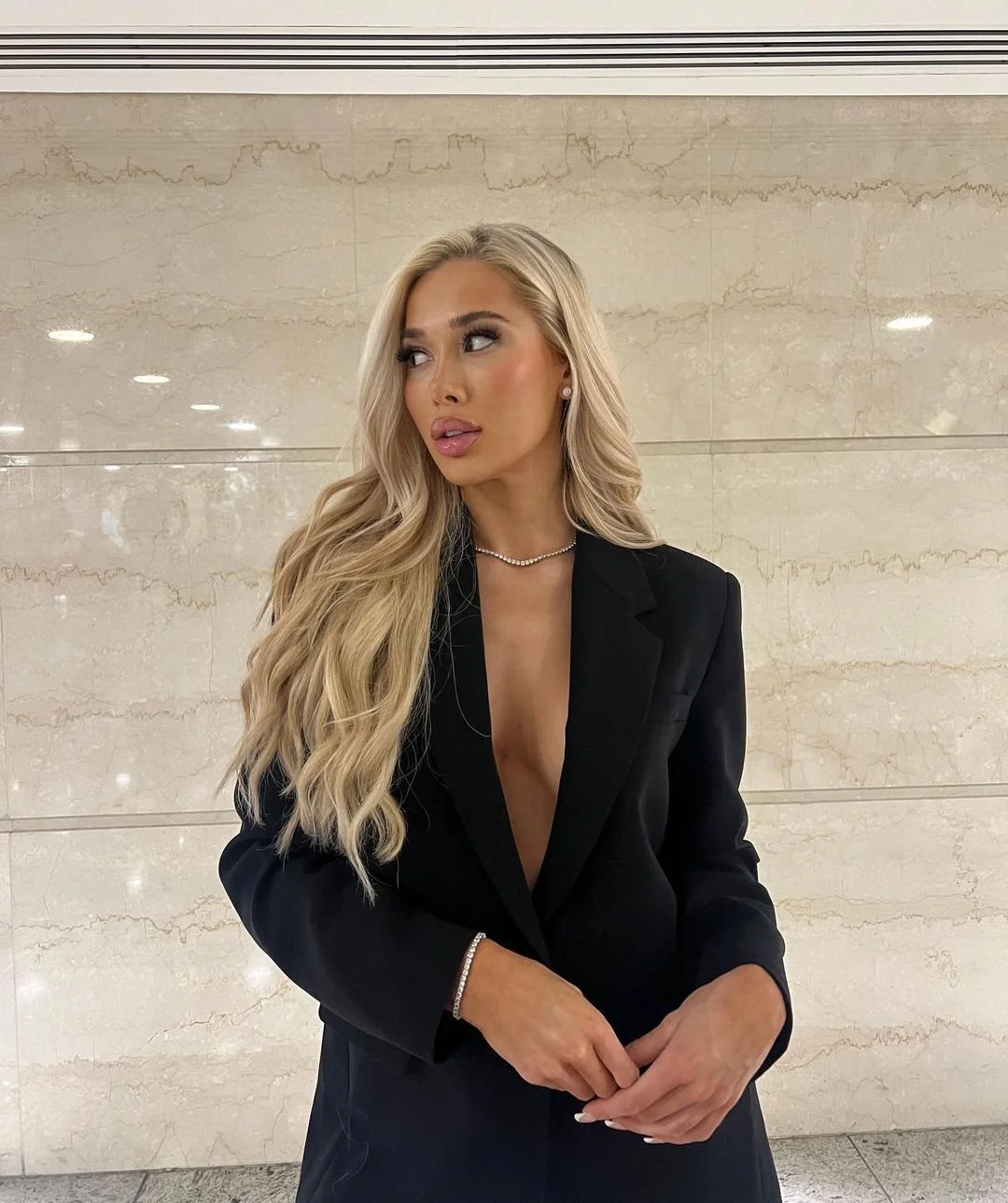 Blonde woman with wavy hair wearing a black blazer with a deep neckline, pearl necklace, and earrings, standing against a beige marble wall.